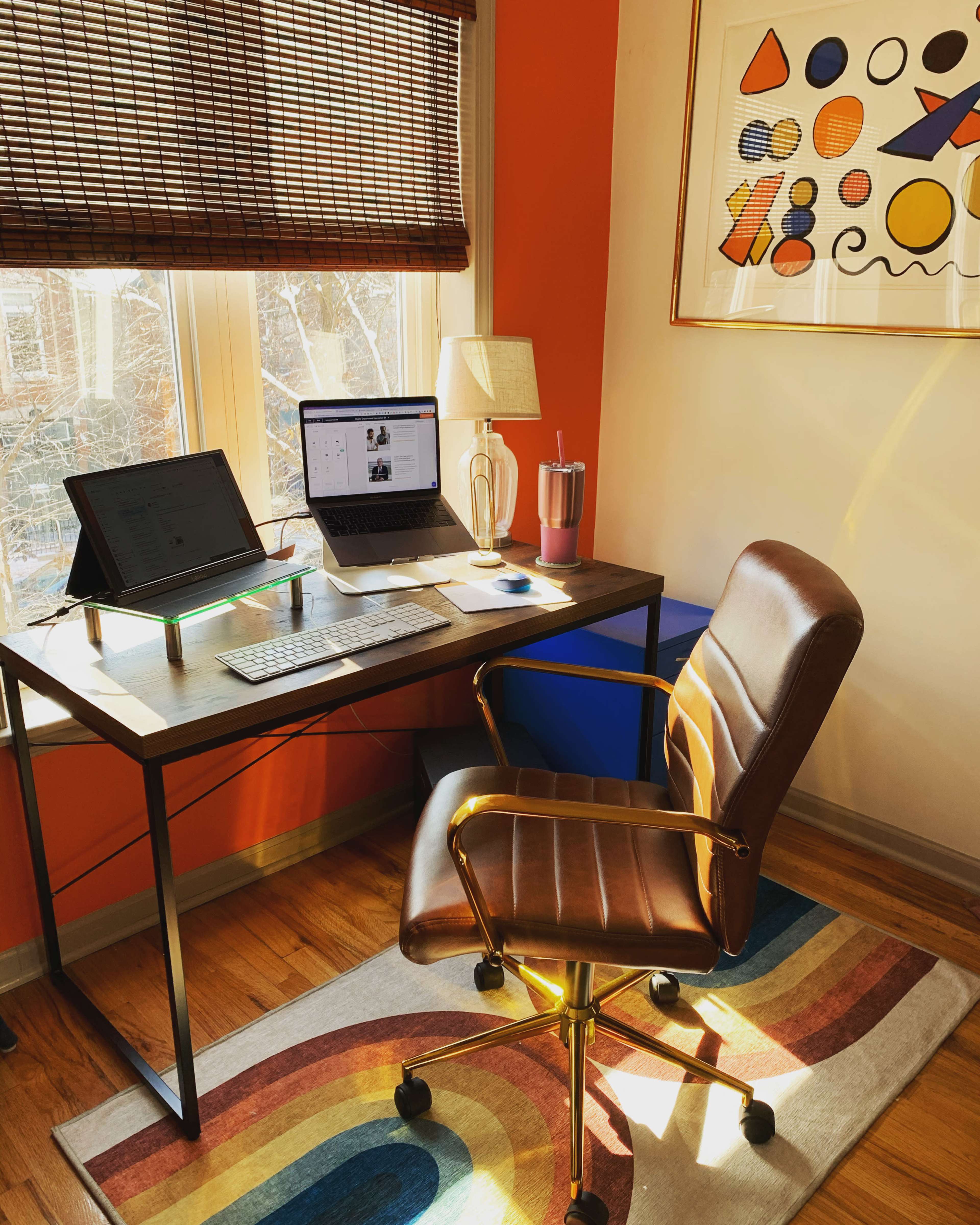 A home office setup features a desk with a laptop, tablet, and accessories, along with a brown office chair and a colorful rug.