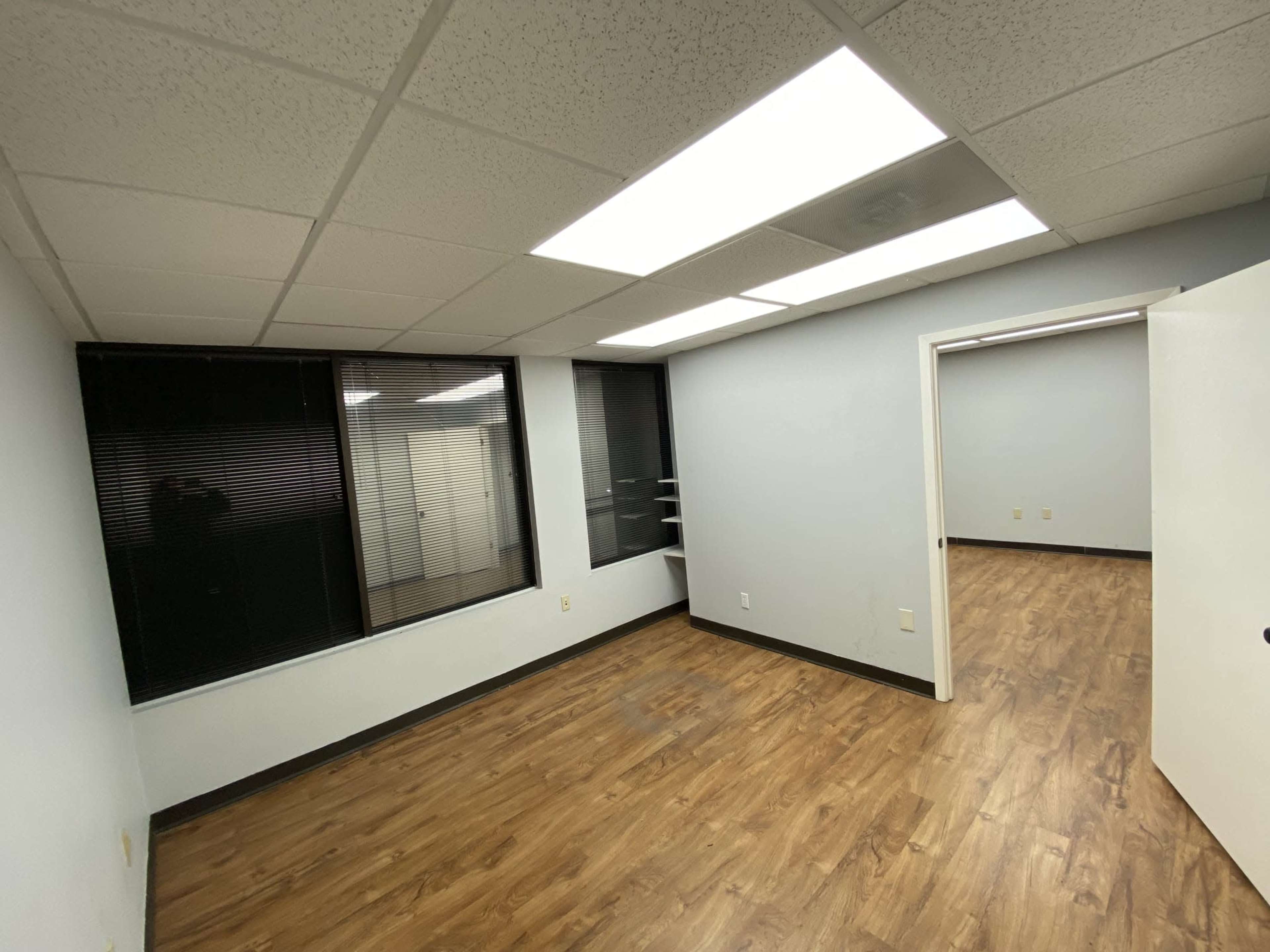 The image shows an empty office space with wooden flooring, gray walls, and large windows with blinds.