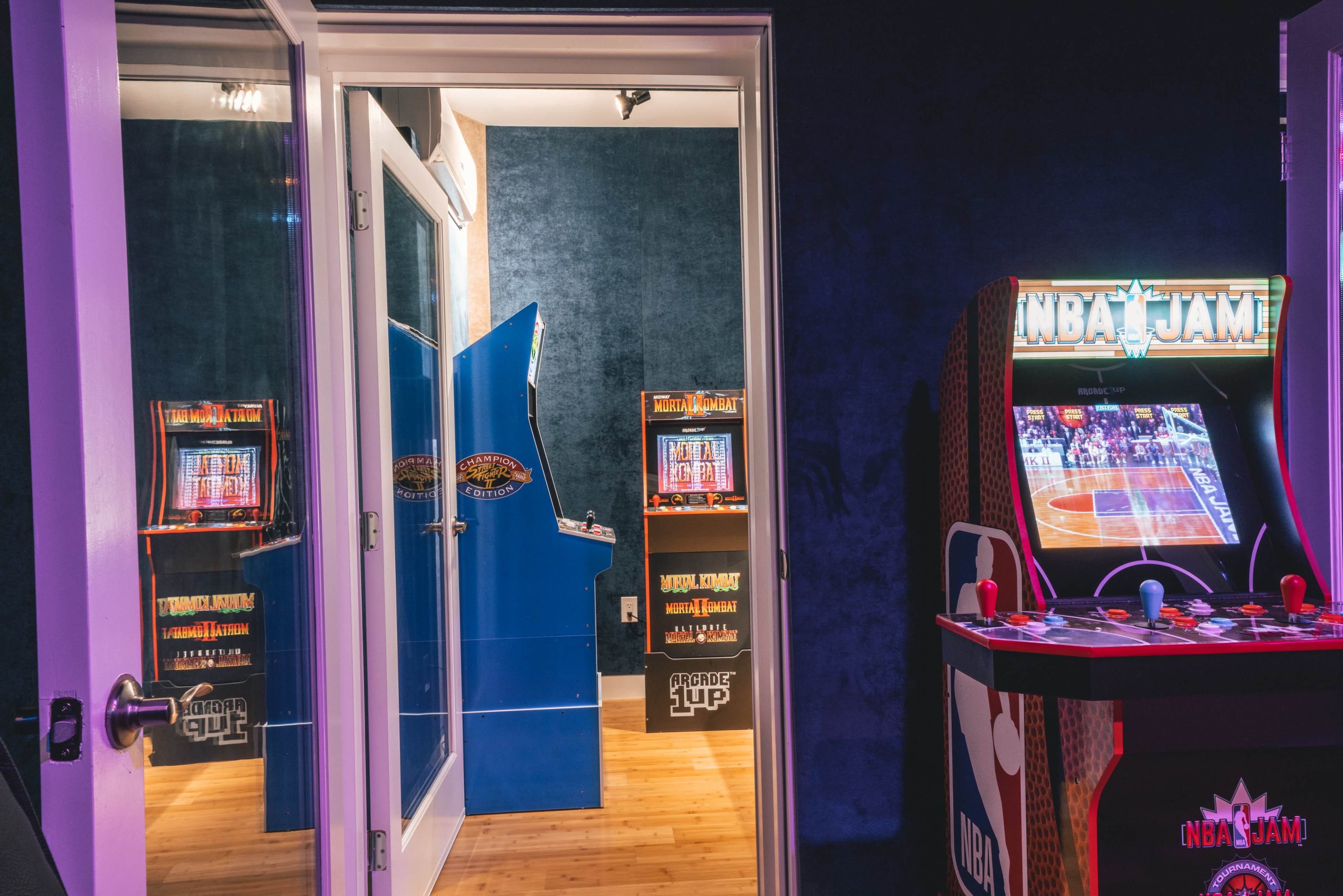 A cozy game room with arcade machines, including "NBA Jam," and a doorway leading to another gaming area.