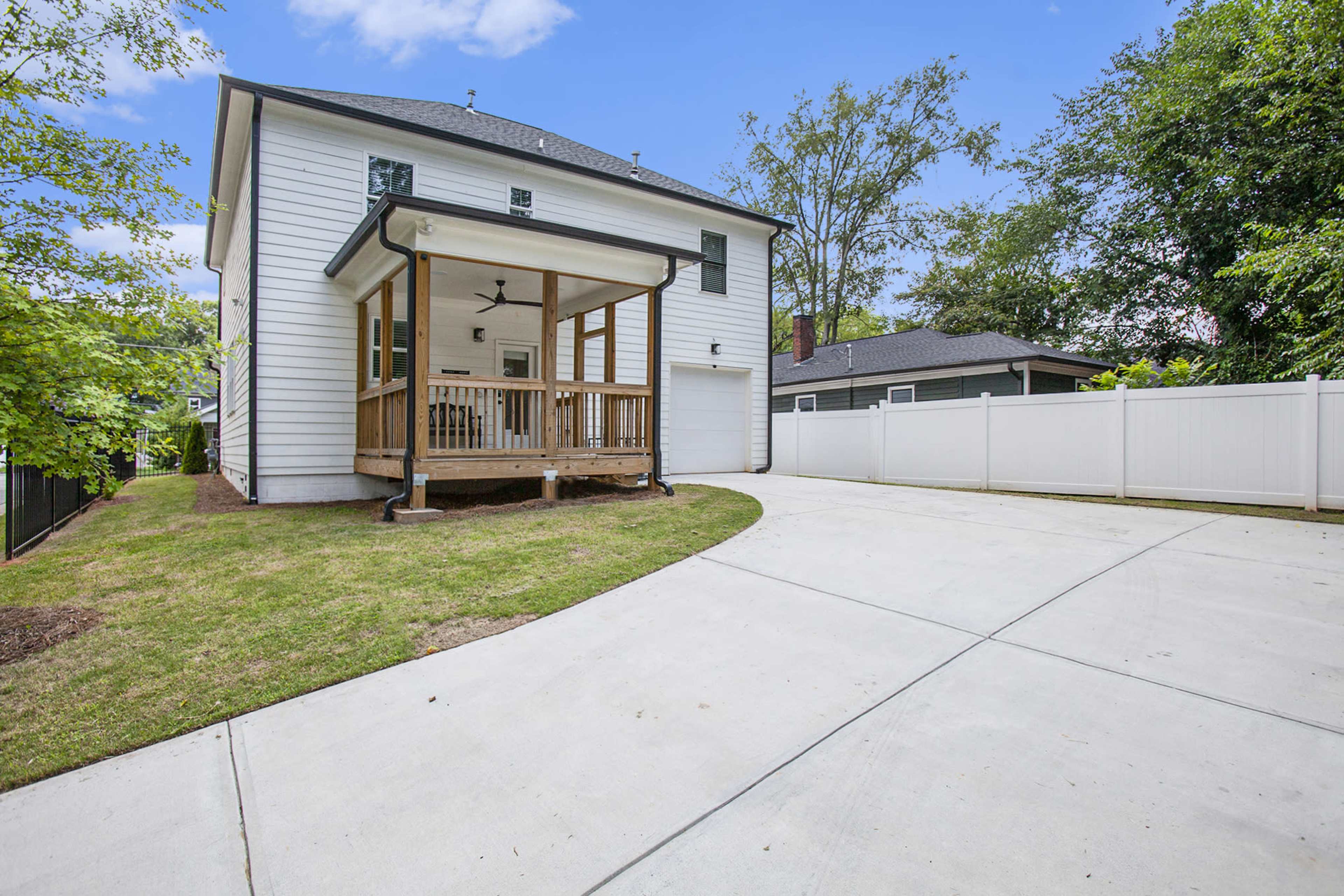 The image shows a two-story white house with a covered front porch, a concrete driveway, and a fenced yard surrounded by trees.