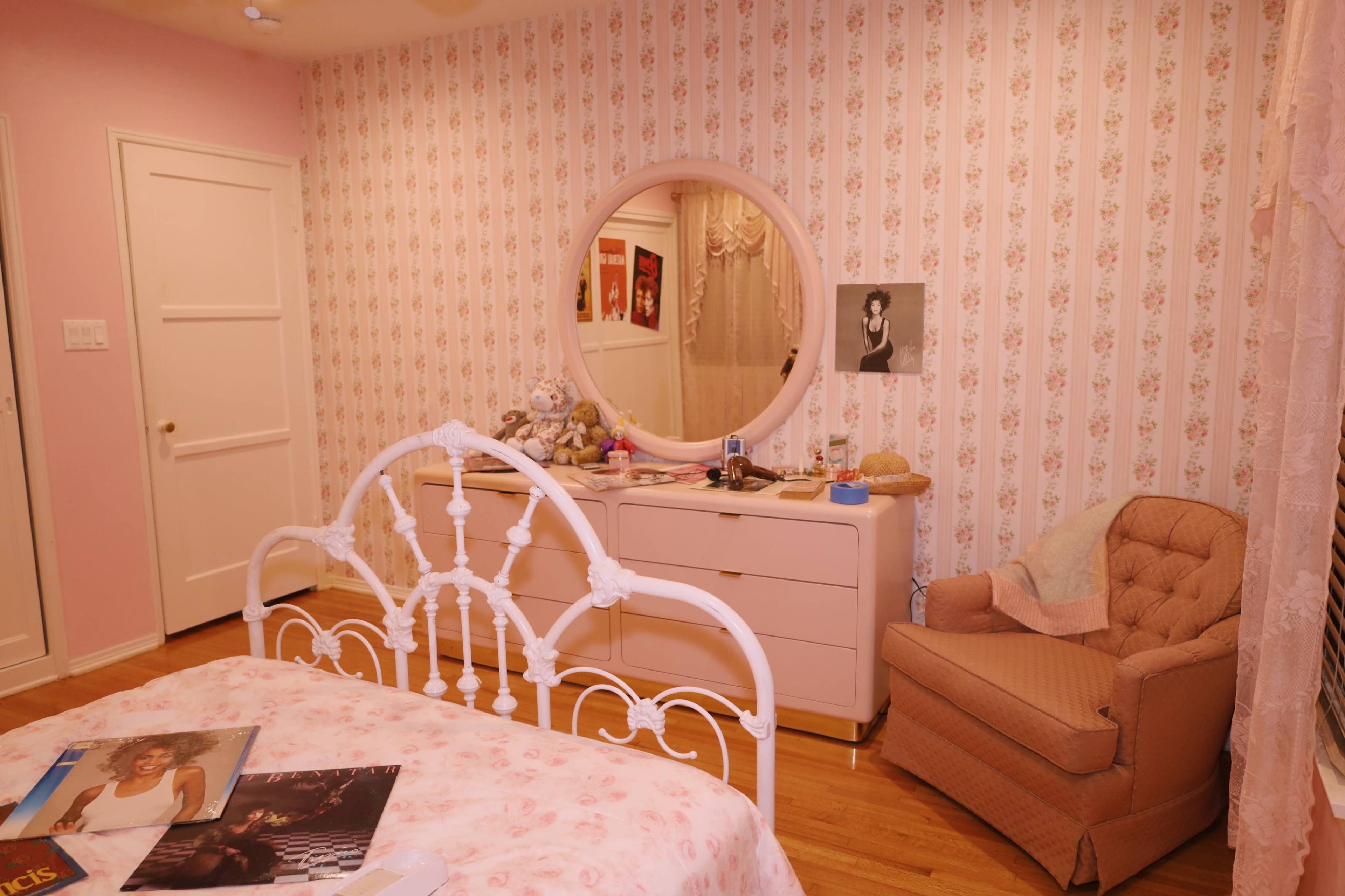 The image shows a bedroom with floral wallpaper, a round mirror above a dresser, and a bed with a pink floral bedspread, accompanied by a chair in the corner.
