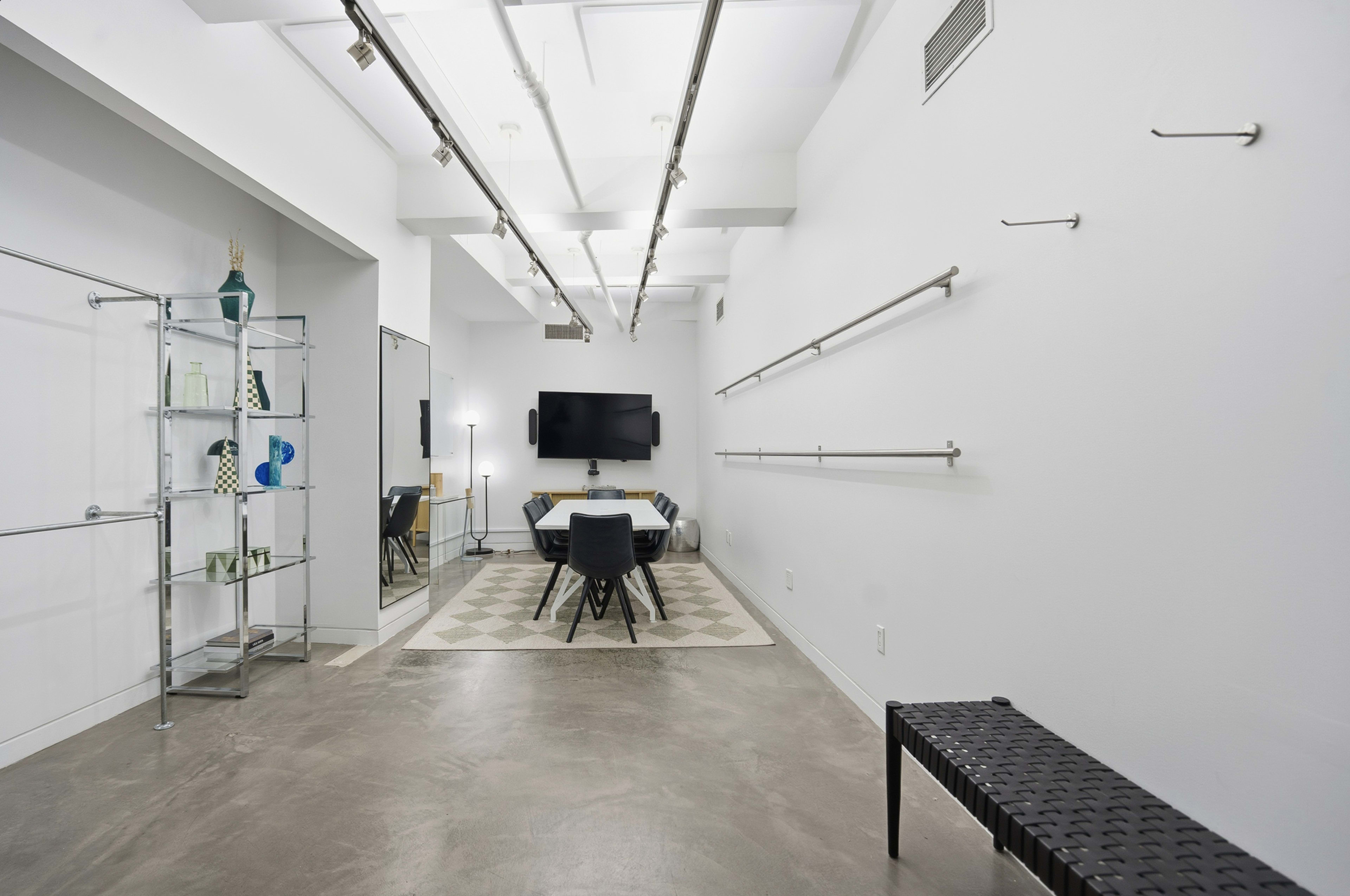 The image shows a modern, minimalist room featuring a large table with chairs, a wall-mounted television, decorative shelves with glass items, and various lighting fixtures.