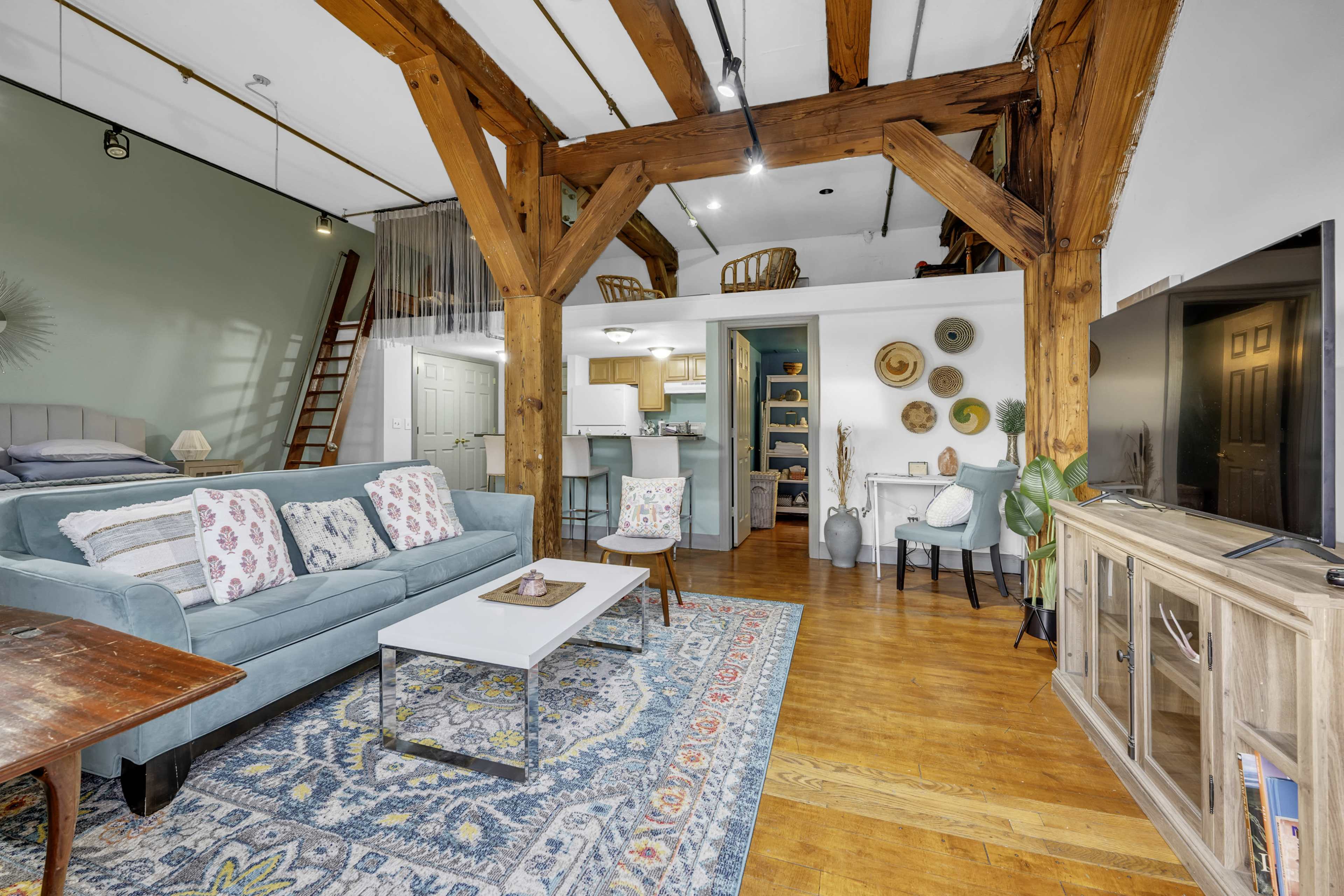 The image shows a spacious living area featuring wooden beams, a blue couch, a coffee table, and a decorative rug, with a staircase leading to an upper level.