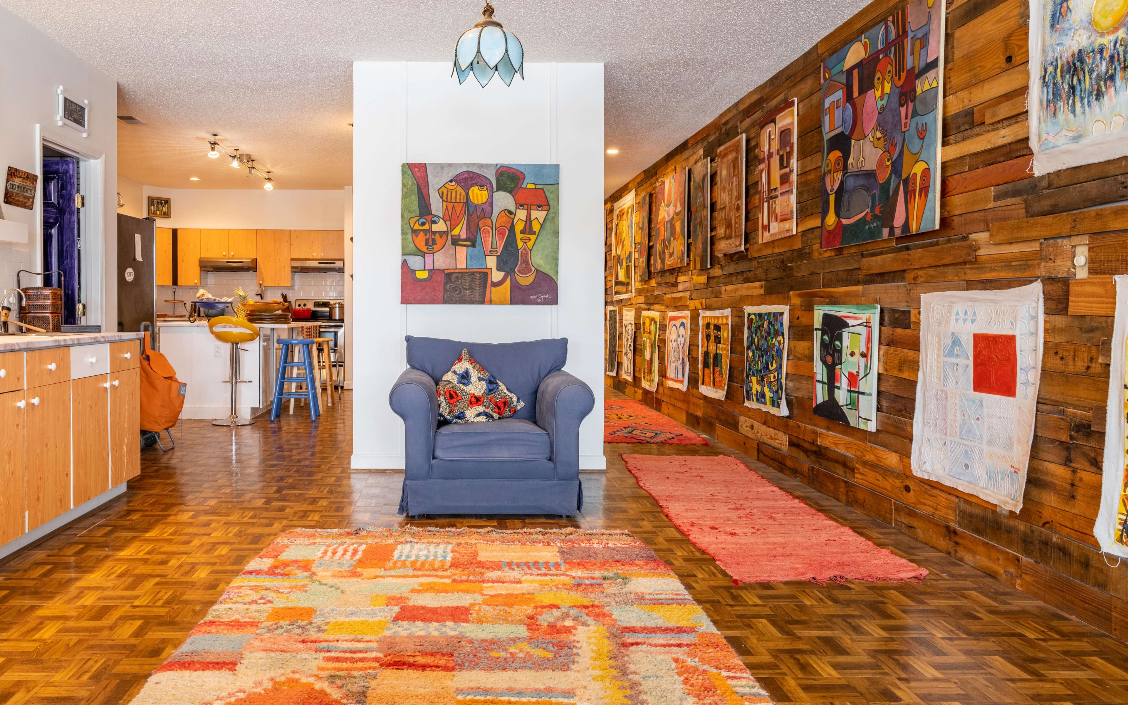 An inviting living room features a blue chair, wooden accent walls adorned with colorful artwork, and a patterned rug on a parquet floor.