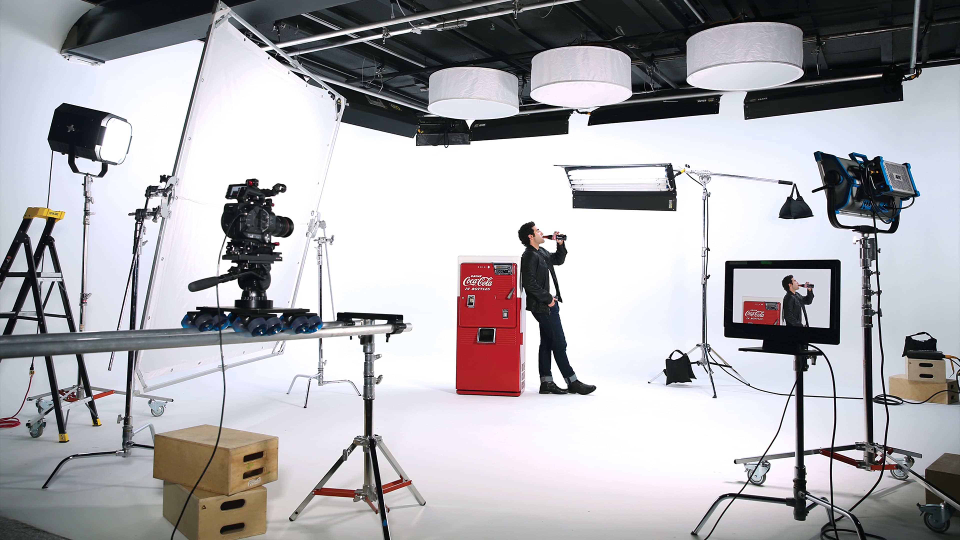 A man in a black suit stands by a red Coca-Cola vending machine in a photo studio filled with lighting equipment and backdrops.
