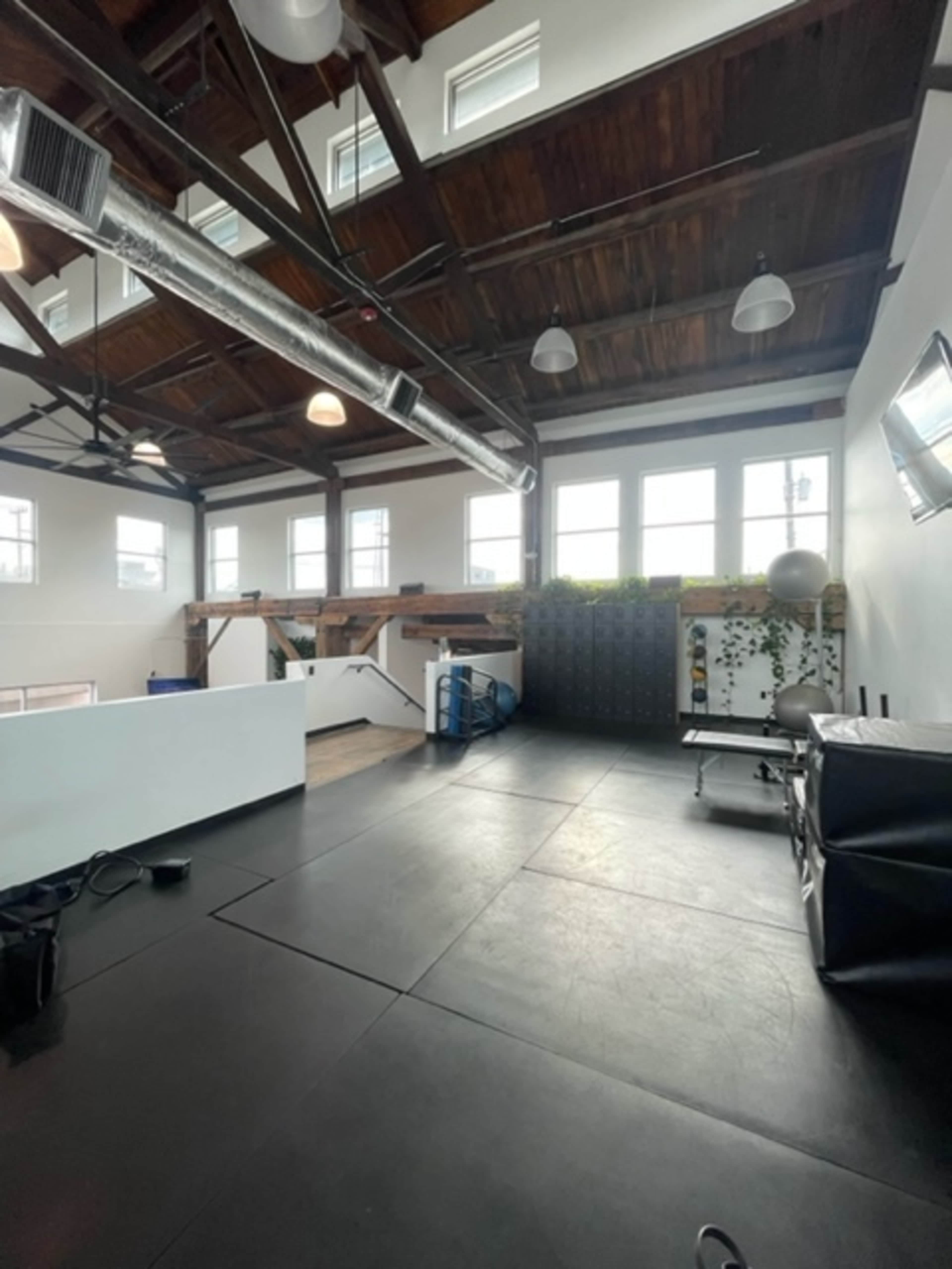 The image shows an open gym space with high wooden ceilings, large windows, and various fitness equipment arranged along the walls.