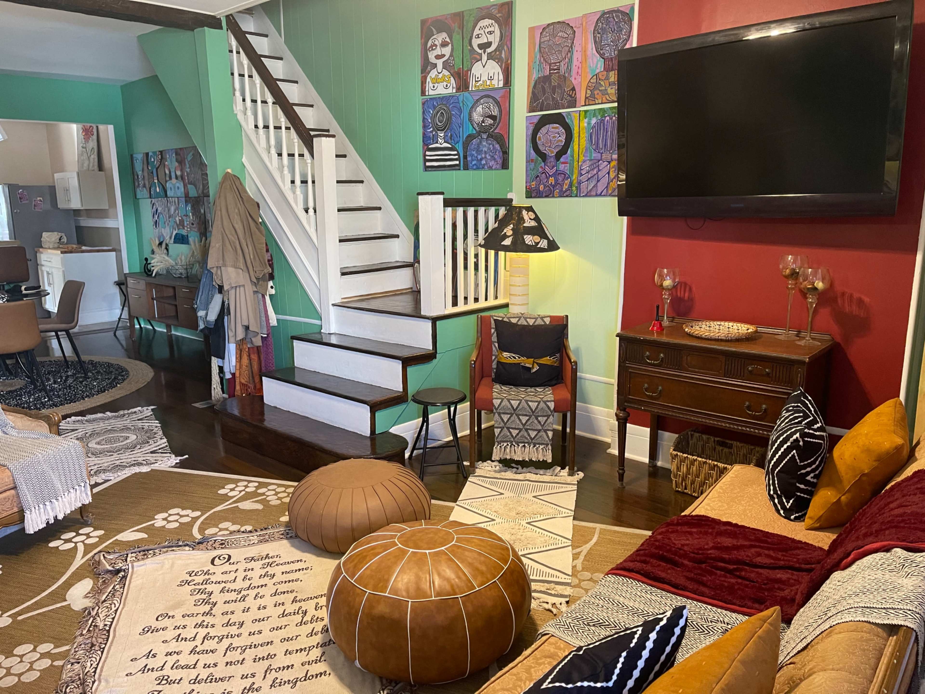 The image shows a cozy living area with a staircase, various decorative artworks on the walls, a television, a couch with colorful cushions, and a patterned area rug.