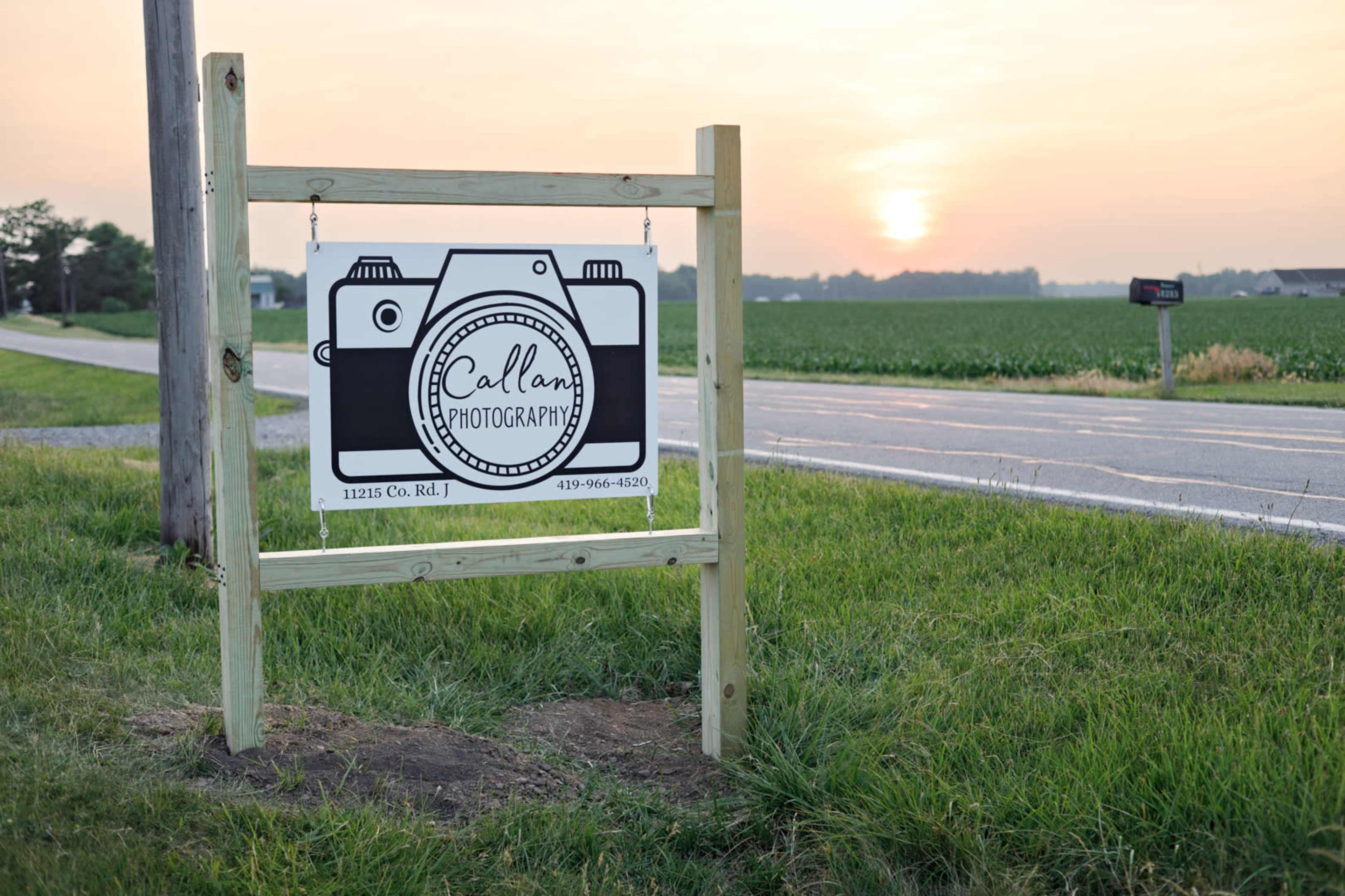 A wooden sign for Callan Photography is posted along a rural road, with a small camera graphic and a sunset in the background.