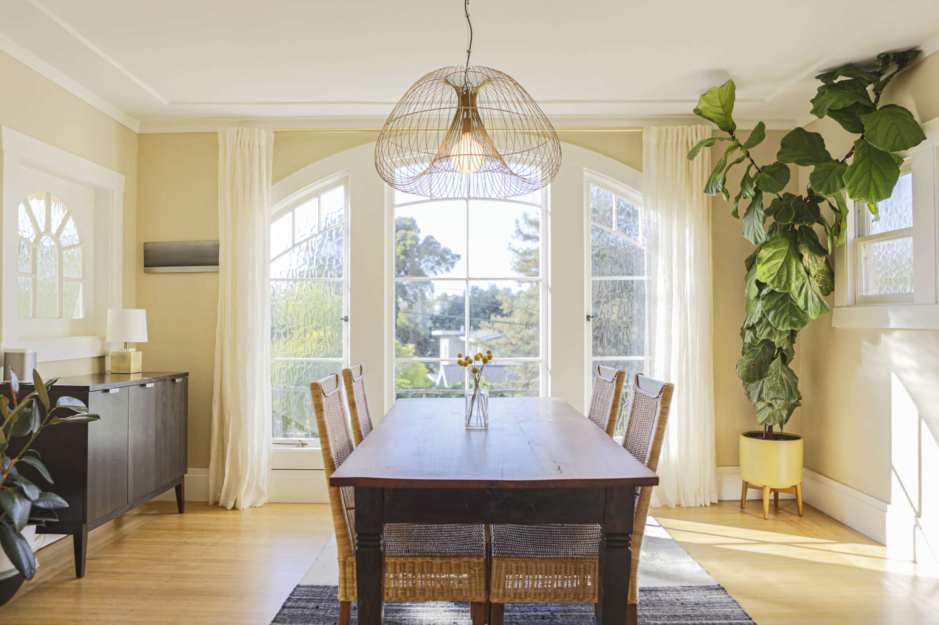 A wooden dining table is centered in a light-filled room with large windows and a plant in the corner.