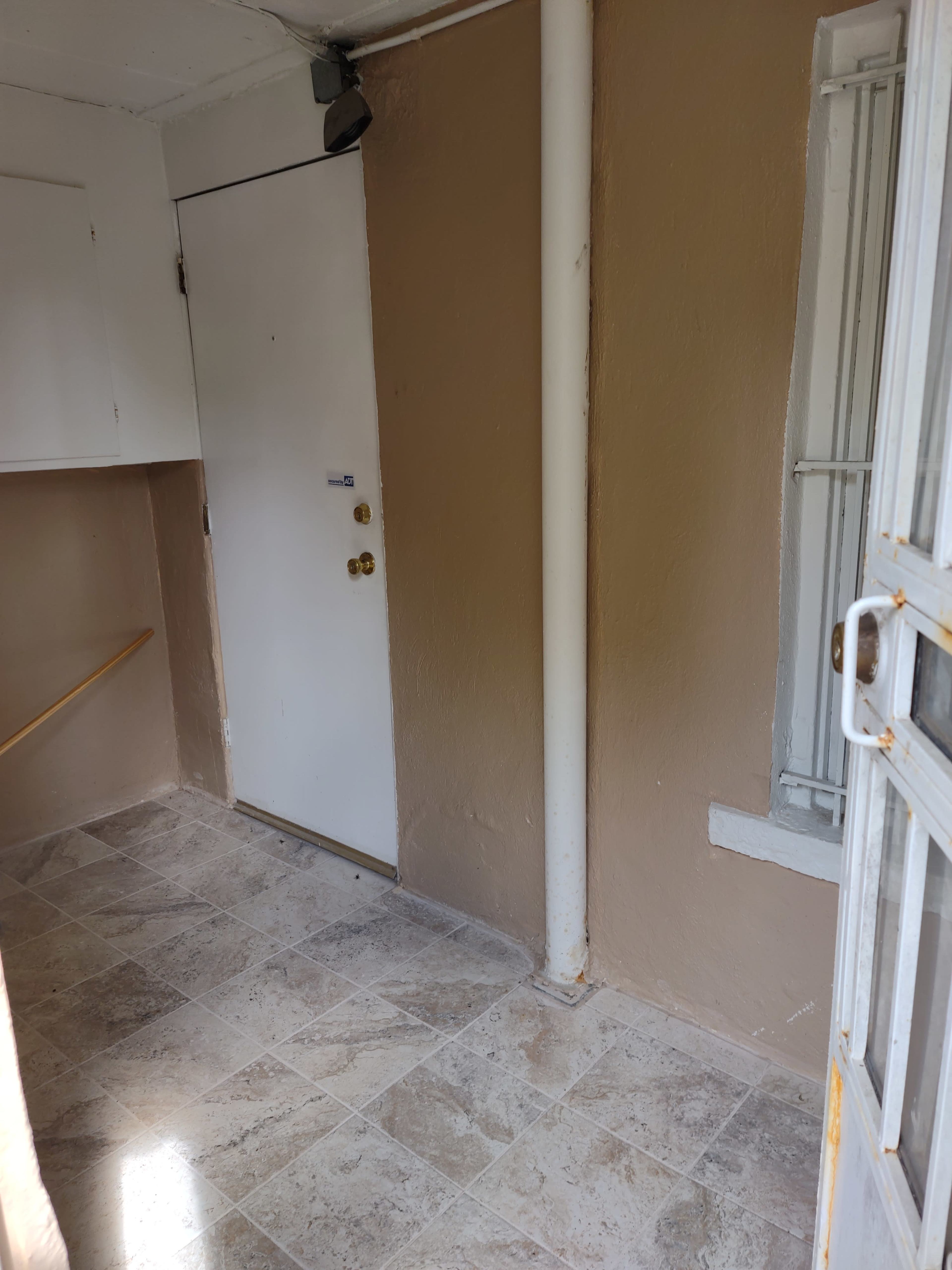 A small, empty entryway with a white door and beige walls, featuring a tiled floor and a window with metal bars.