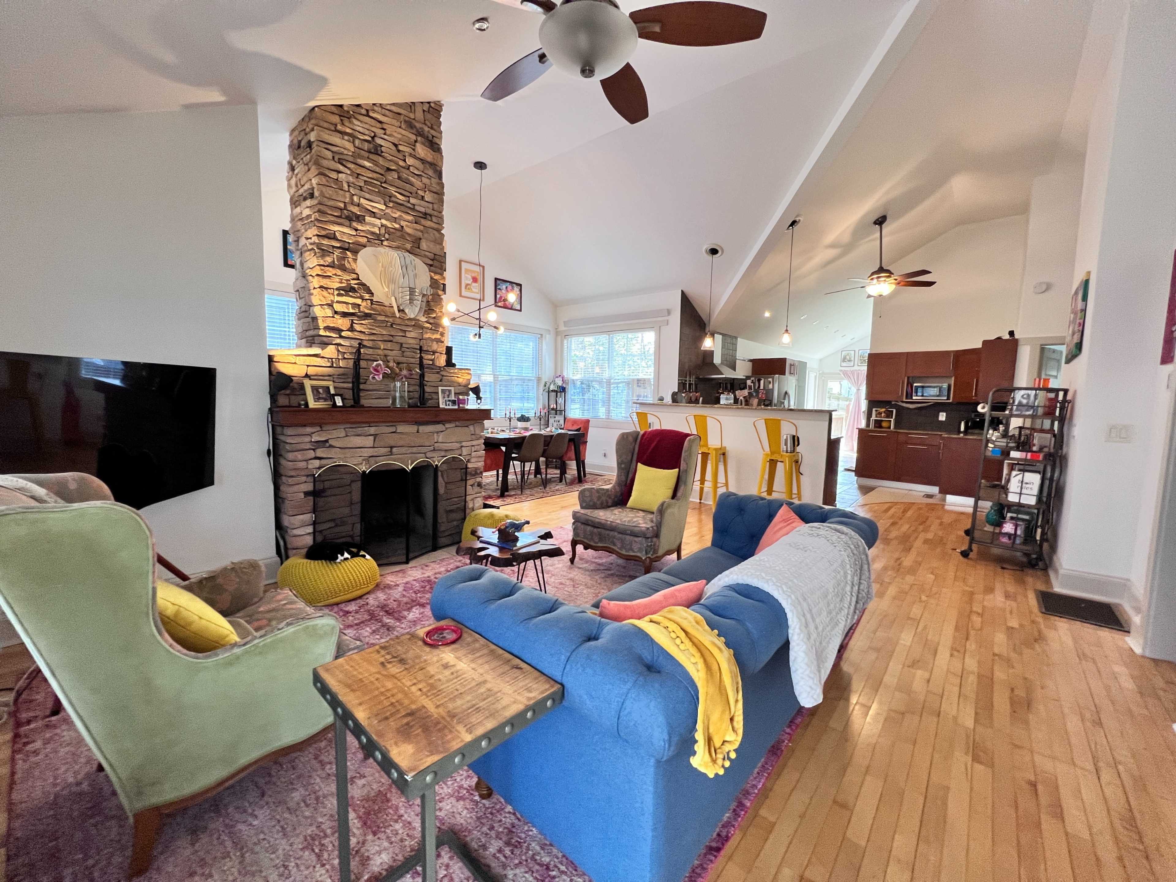 The image shows a living room with a stone fireplace, colorful furniture, and an open layout connecting to a dining area and kitchen.