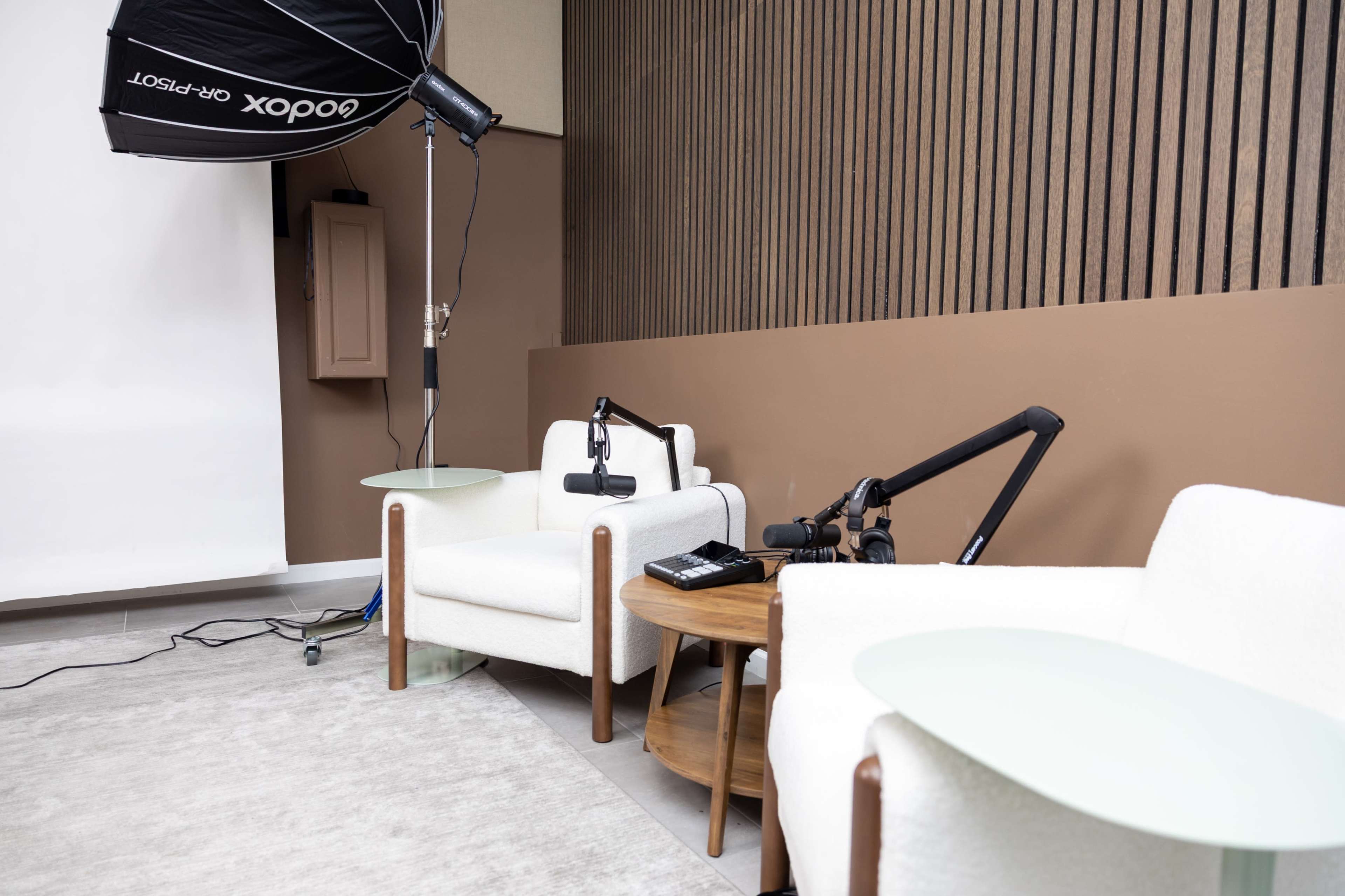 The image features a studio setting with two modern armchairs, green side tables, and an overhead lighting setup against a textured brown wall.