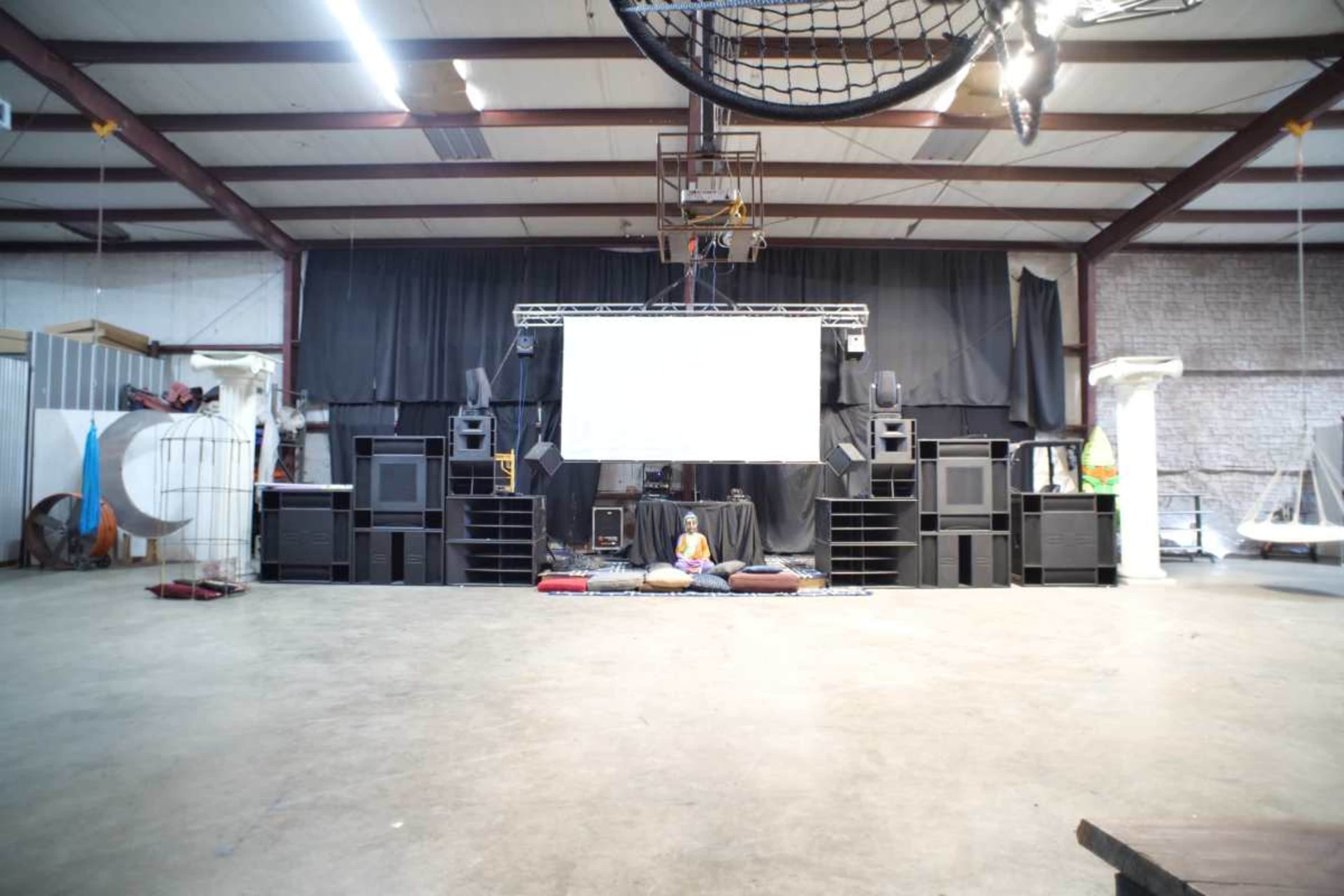 A person sits on cushions in the center of an empty warehouse space with large speakers and a screen in the background.