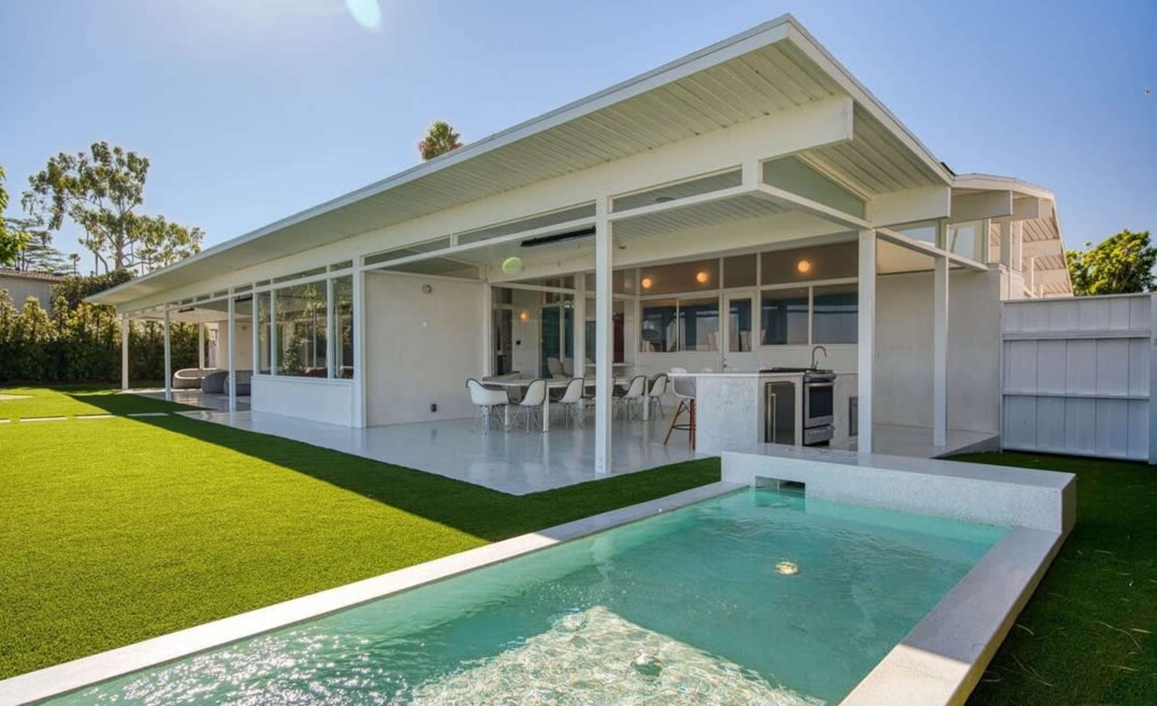 A modern white house with a flat roof features a spacious backyard, an outdoor seating area, and a shallow swimming pool.