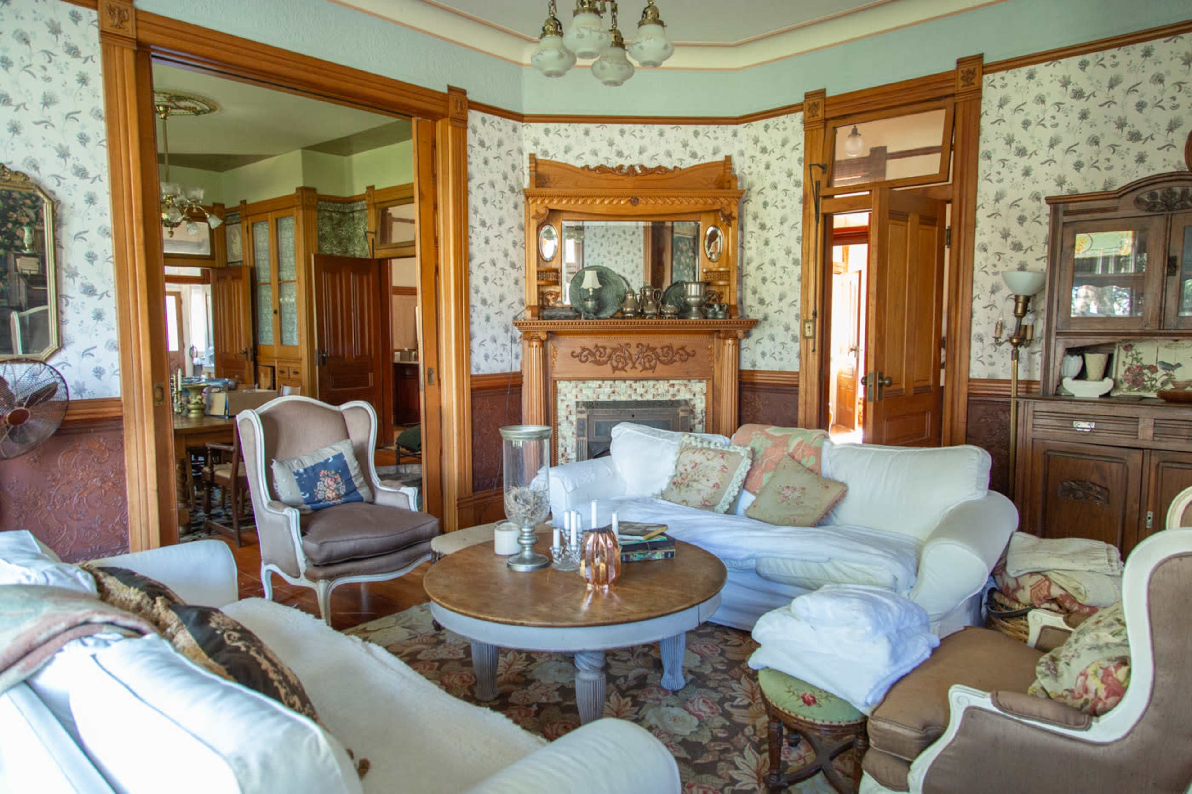 The image shows a vintage living room featuring a circular wooden coffee table surrounded by white upholstered sofas and a large fireplace framed by ornate woodwork.