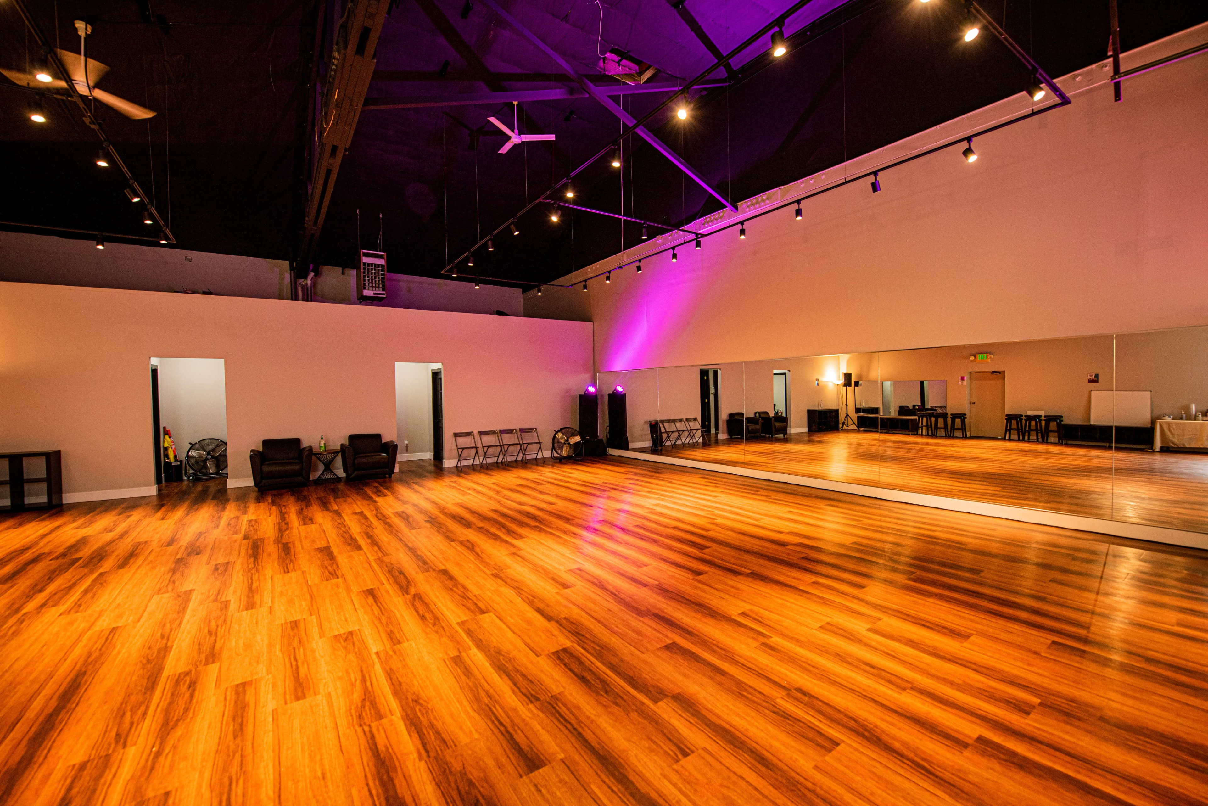 A spacious dance studio with wooden flooring, large mirrors on one wall, and minimal furniture.