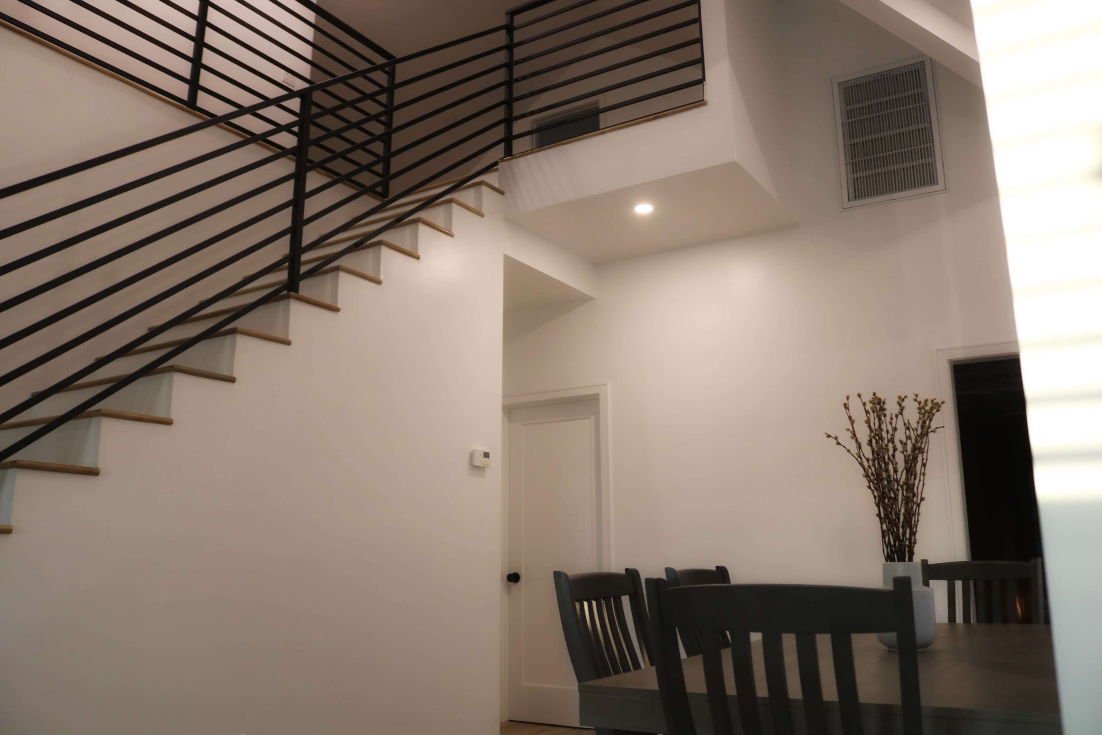 A modern interior with a staircase featuring black railings and a dining area with a table and chairs, illuminated by soft light.
