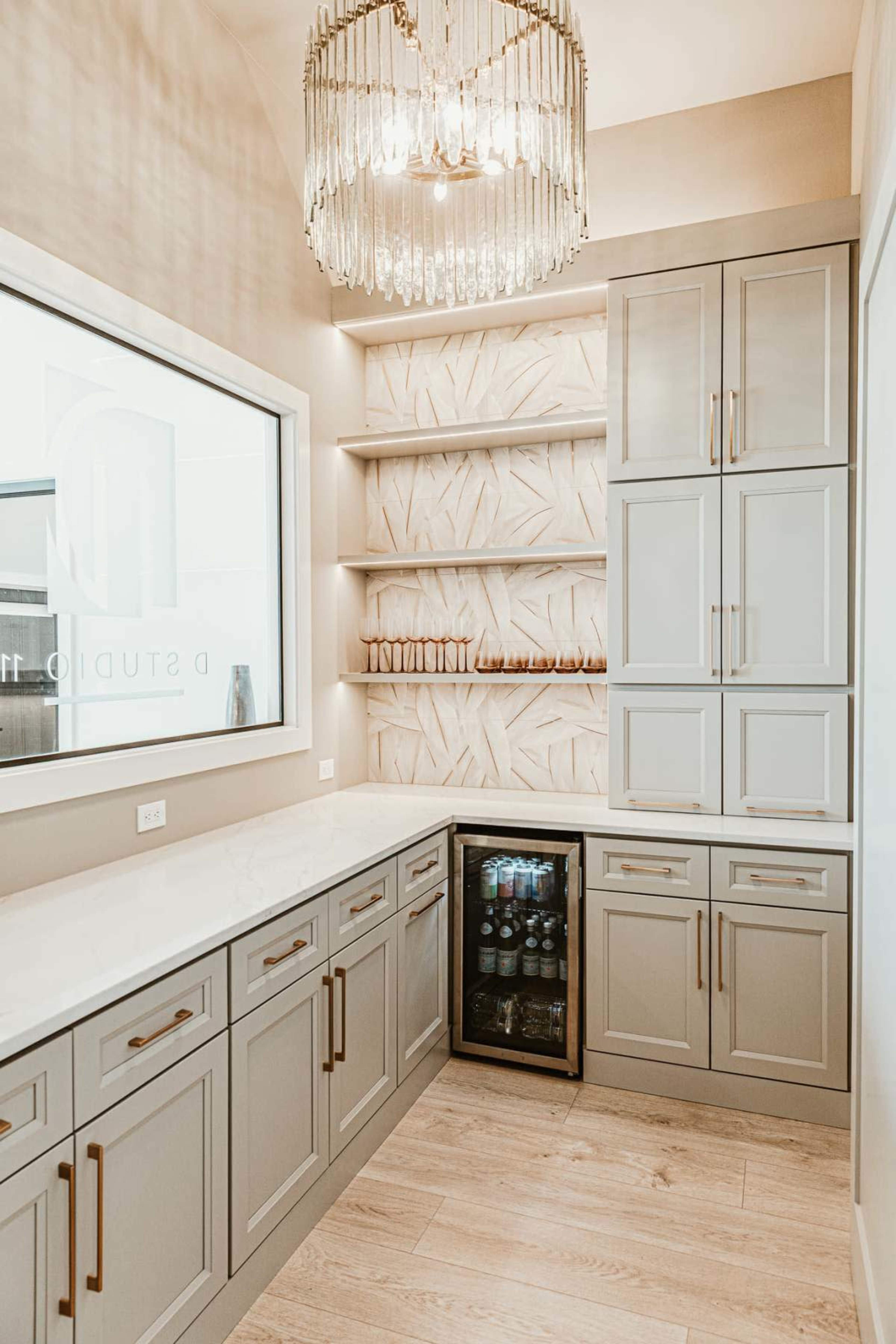 The image shows a modern room with gray cabinetry, a glass-fronted beverage refrigerator, open shelving, and a chandelier illuminated overhead.