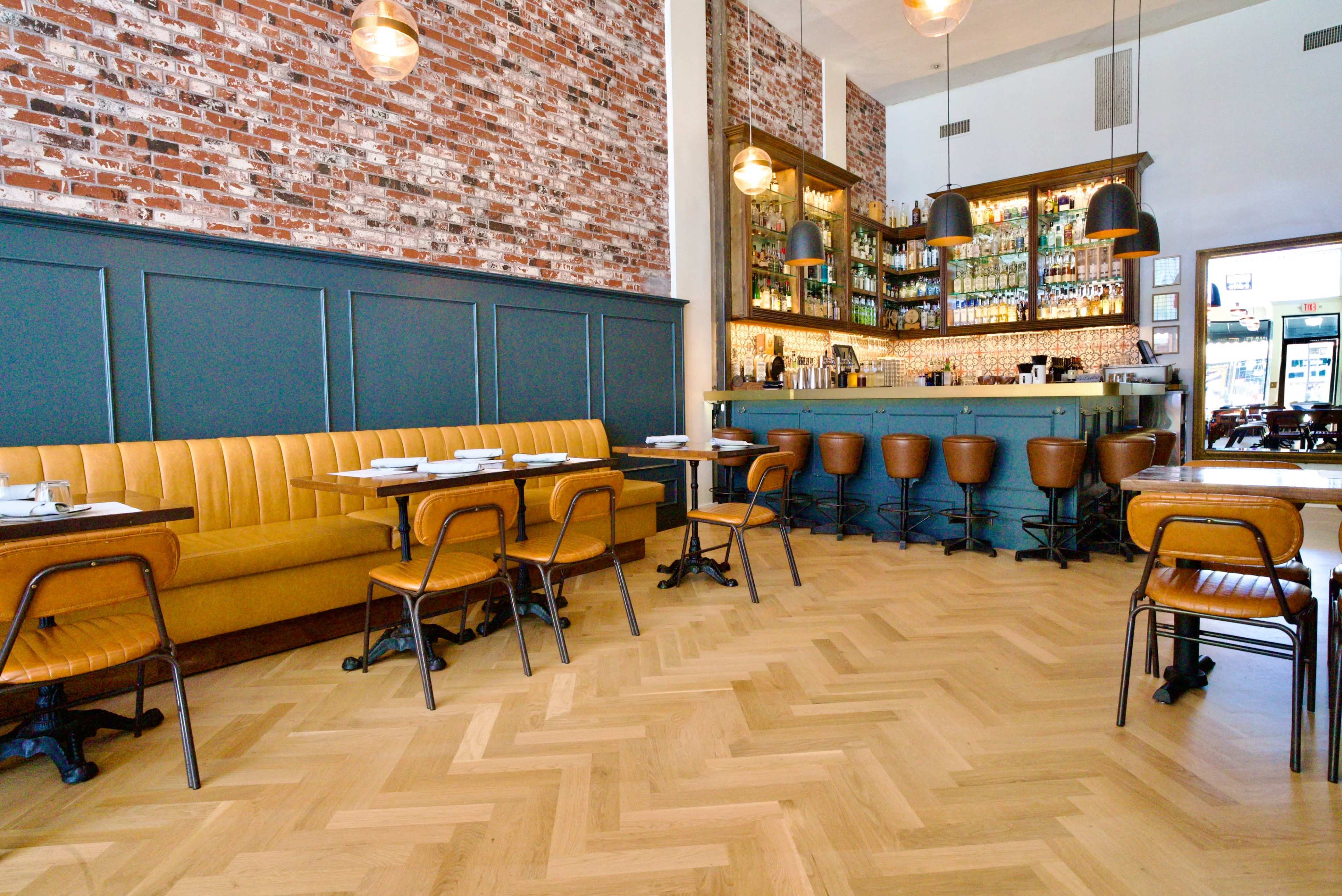 The image shows a modern restaurant interior with a brick wall, a green bar area, and wooden herringbone flooring.