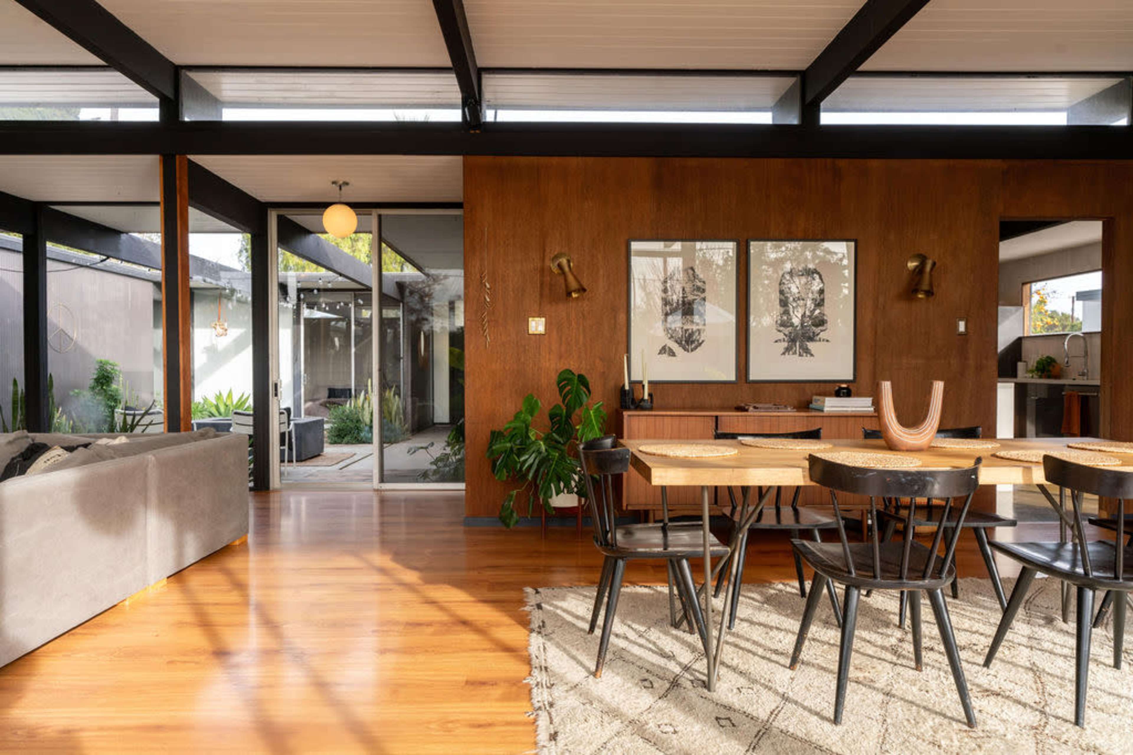 The image shows a modern interior space featuring a dining area with a wooden table and black chairs, surrounded by large windows that provide natural light.