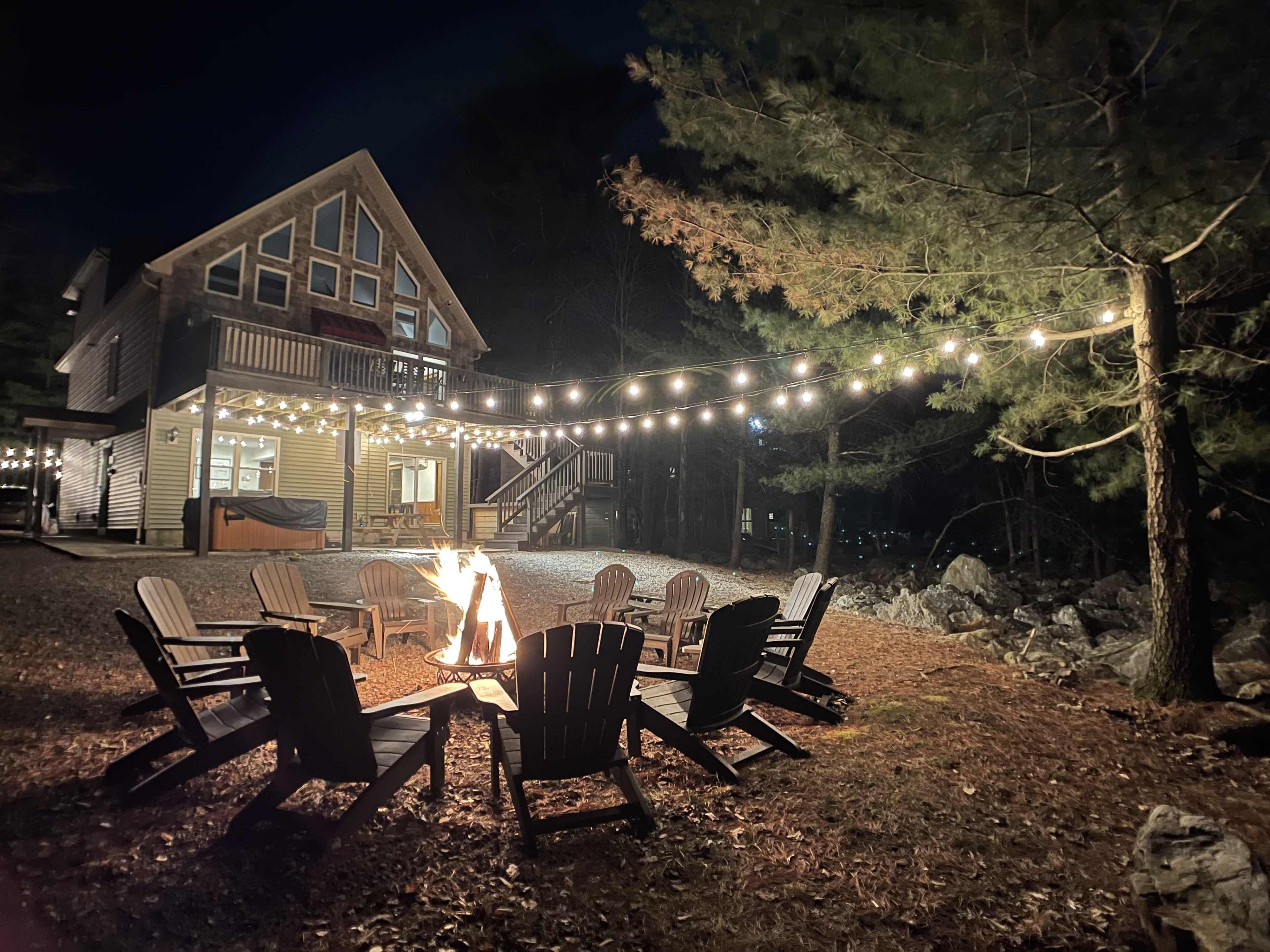 A fire pit surrounded by wooden chairs is set in front of a house illuminated by string lights at night, with trees in the background.