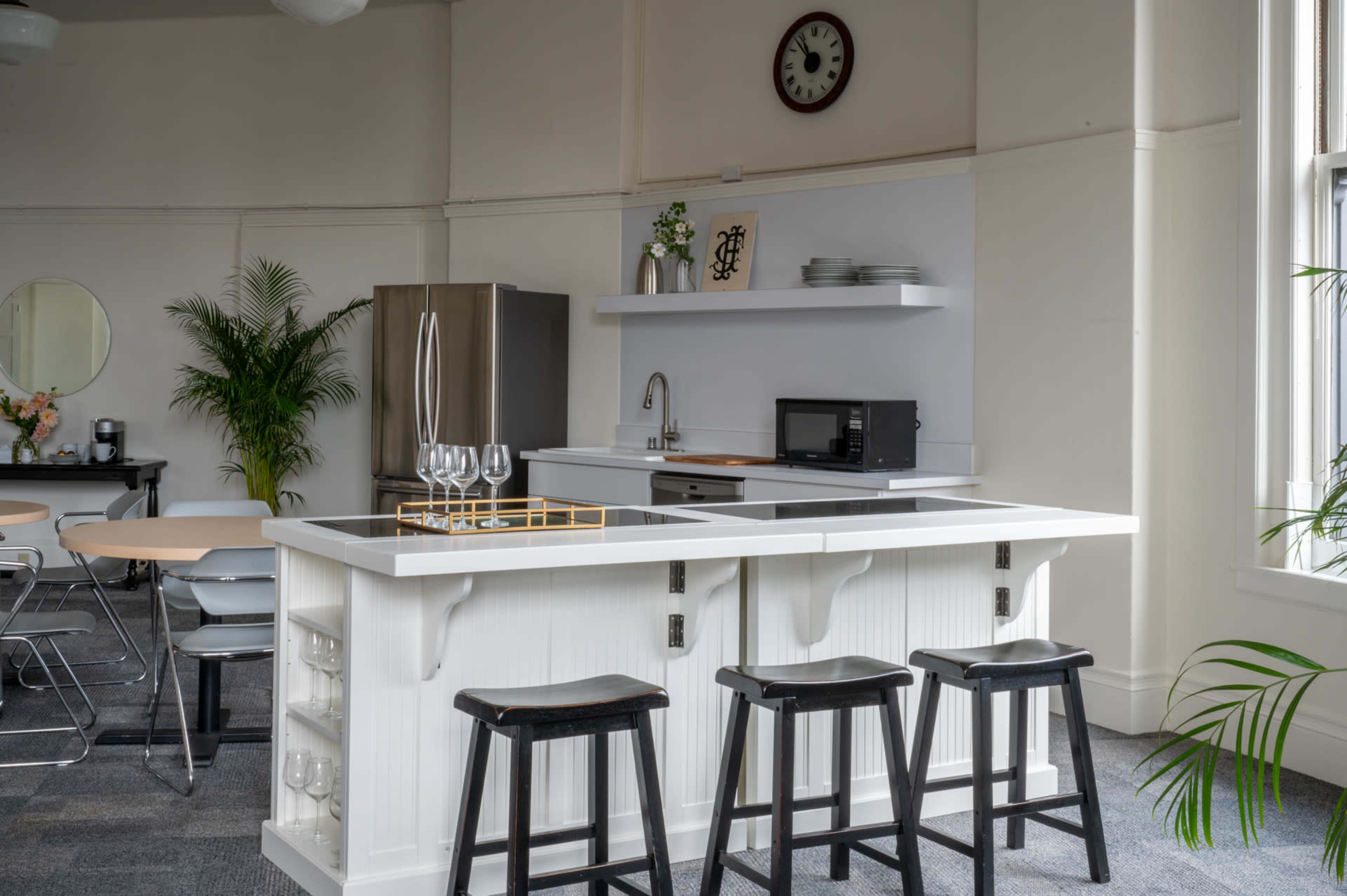 A modern kitchen features a white island with bar stools, a sink, and a microwave, set against a backdrop of a spacious, well-lit room with plants and a clock on the wall.