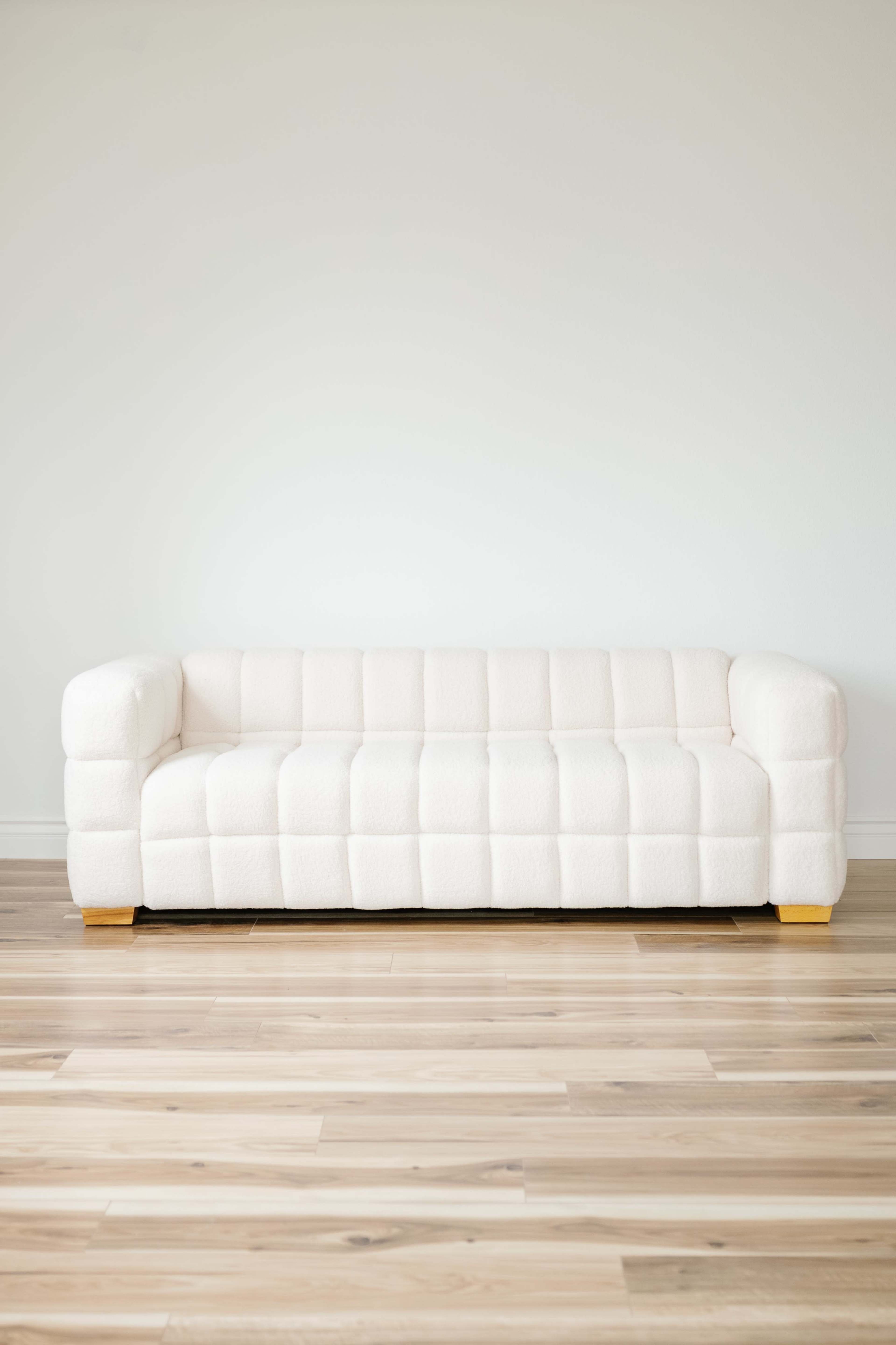 A white, tufted sofa is placed against a plain wall with wooden flooring.