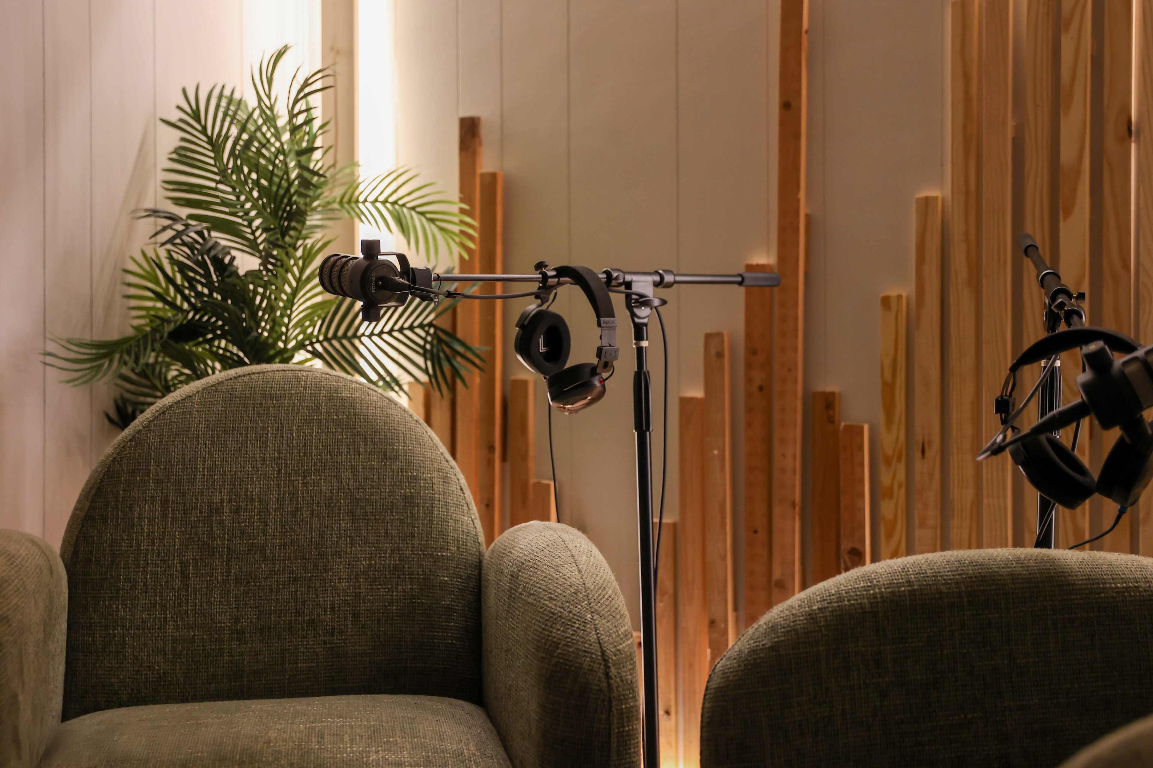 The image depicts two green chairs facing a microphone setup with headphones, surrounded by wooden decor and a plant.