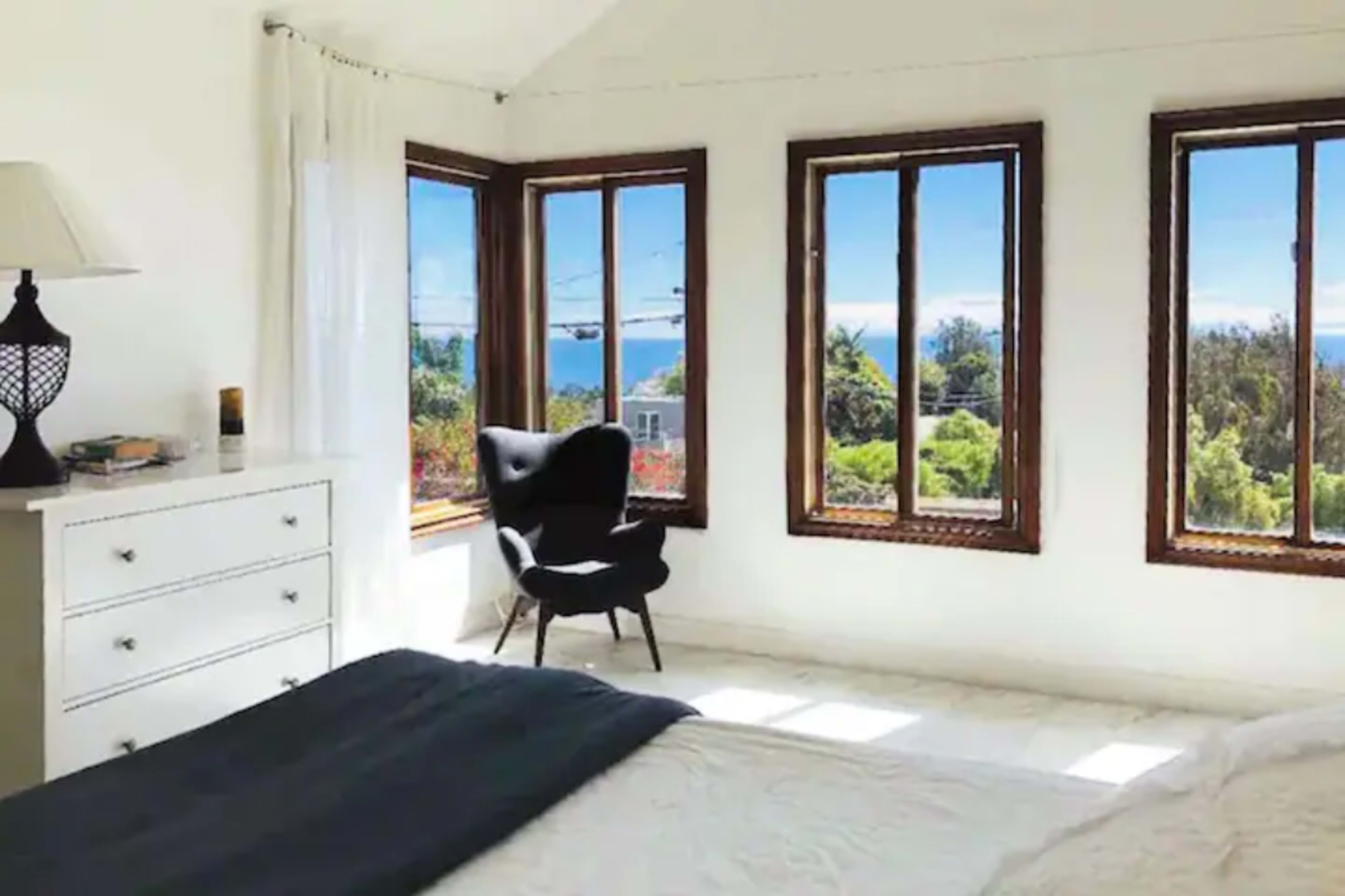 A bright bedroom features a bed with a dark throw and a black chair next to large windows that showcase a view of greenery and the ocean.