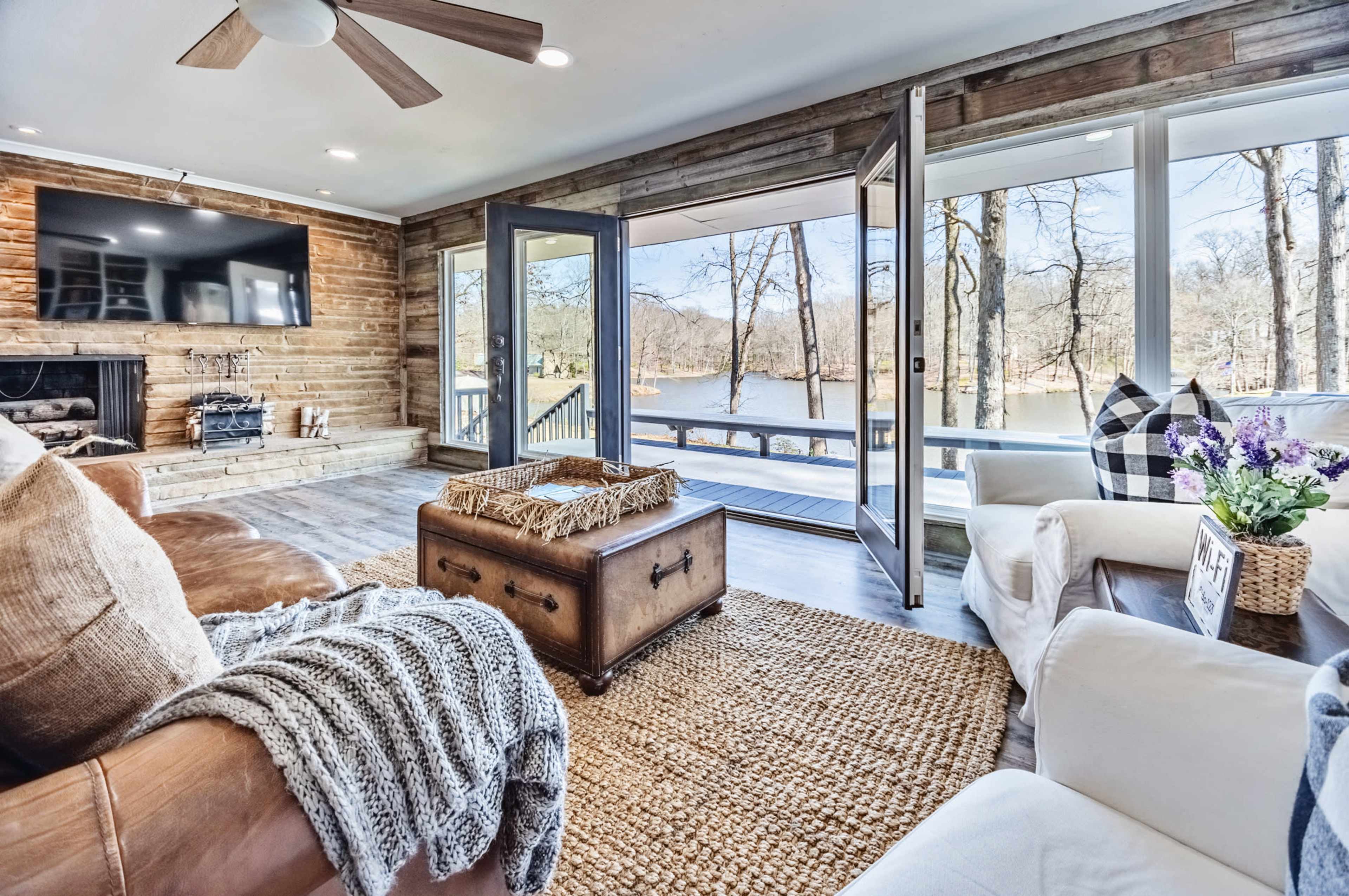 The image shows a cozy living room with large windows overlooking a lake, featuring wooden walls, comfortable seating, and a coffee table.