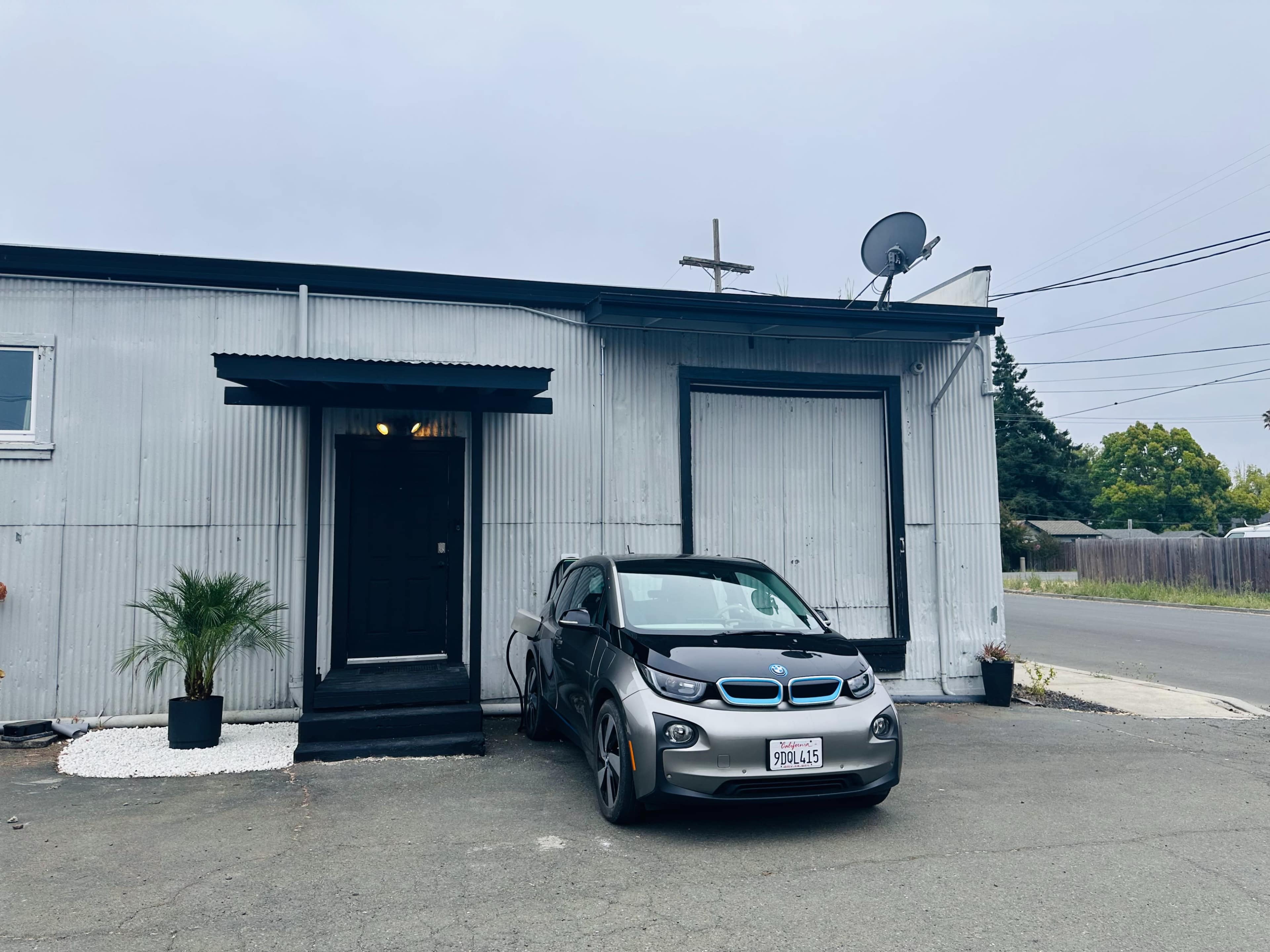 A grey BMW i3 is parked in front of a corrugated metal building with a black door and a satellite dish.