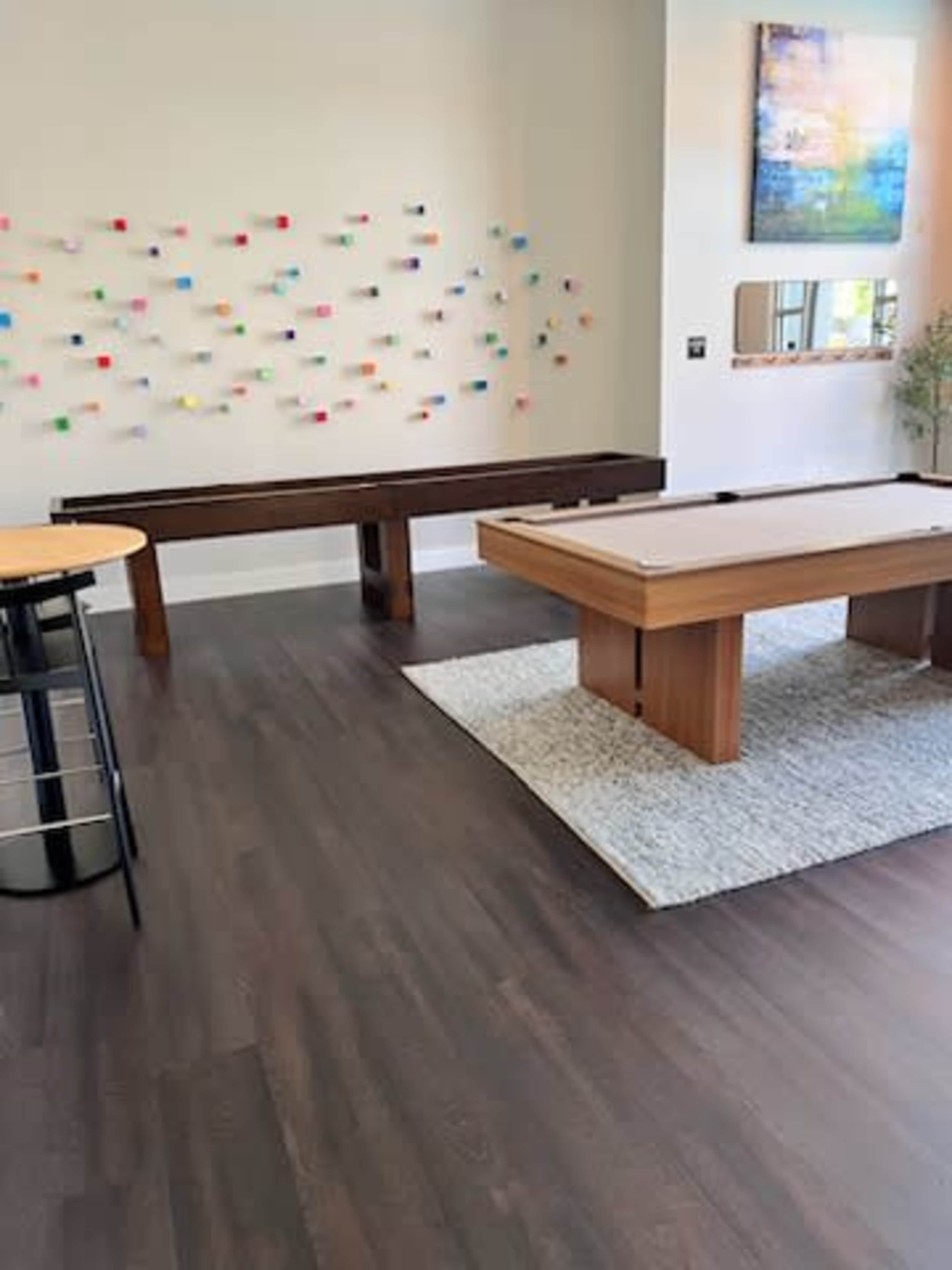 A modern game room with a shuffleboard table, a wooden bench against a colorful wall installation, and a large window letting in natural light.