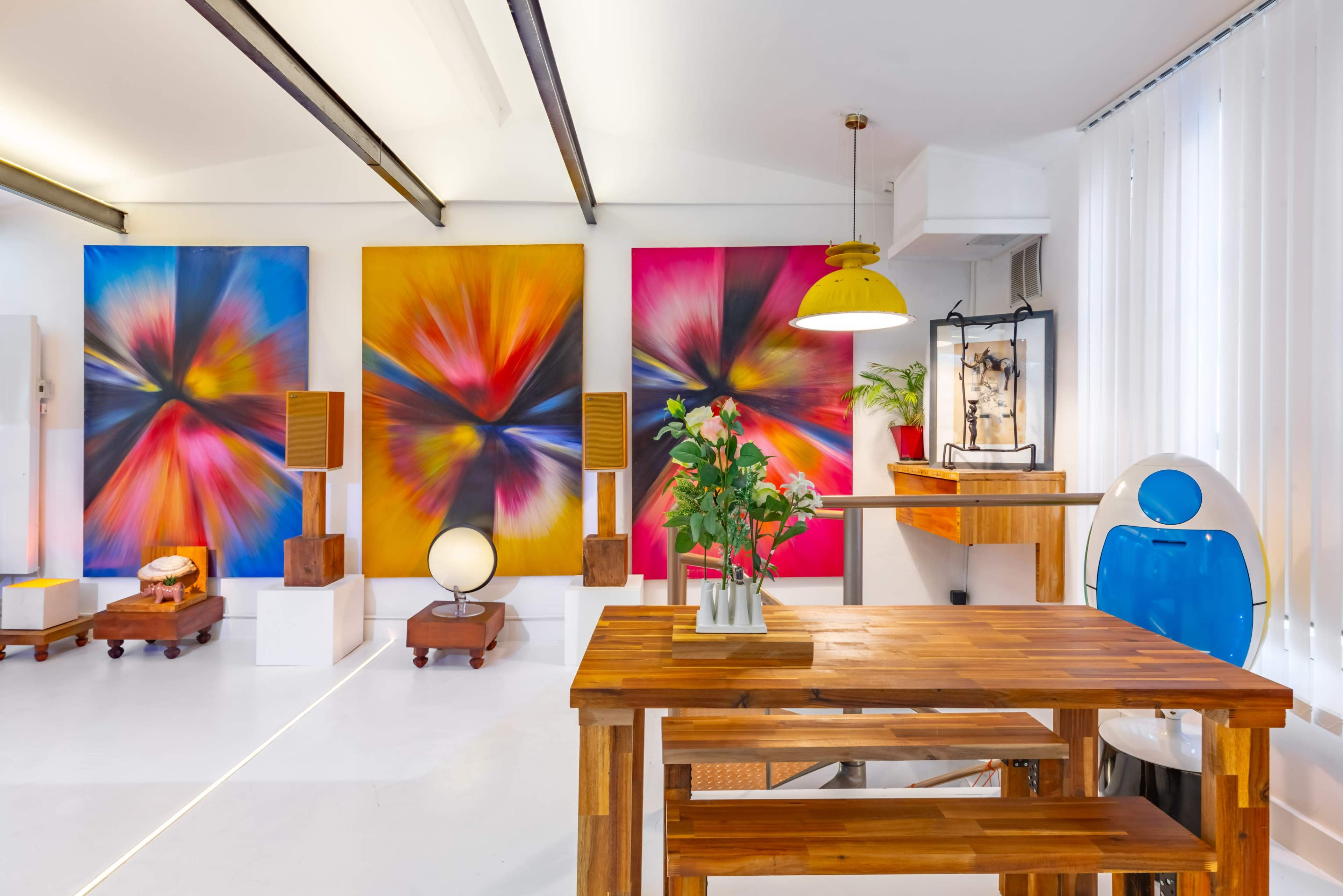 A well-lit art studio features a wooden table surrounded by colorful abstract paintings, a globe, and modern decor.