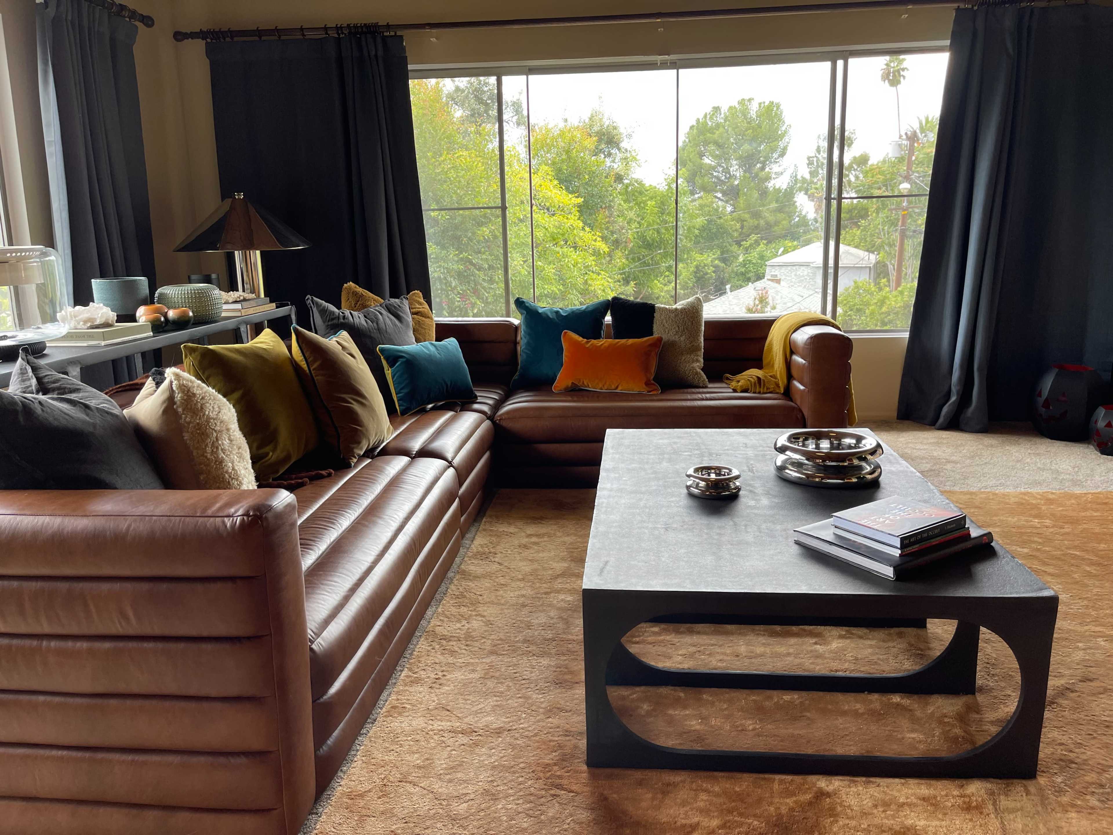 A cozy living room with a brown leather couch, a coffee table, and large windows overlooking a green garden.