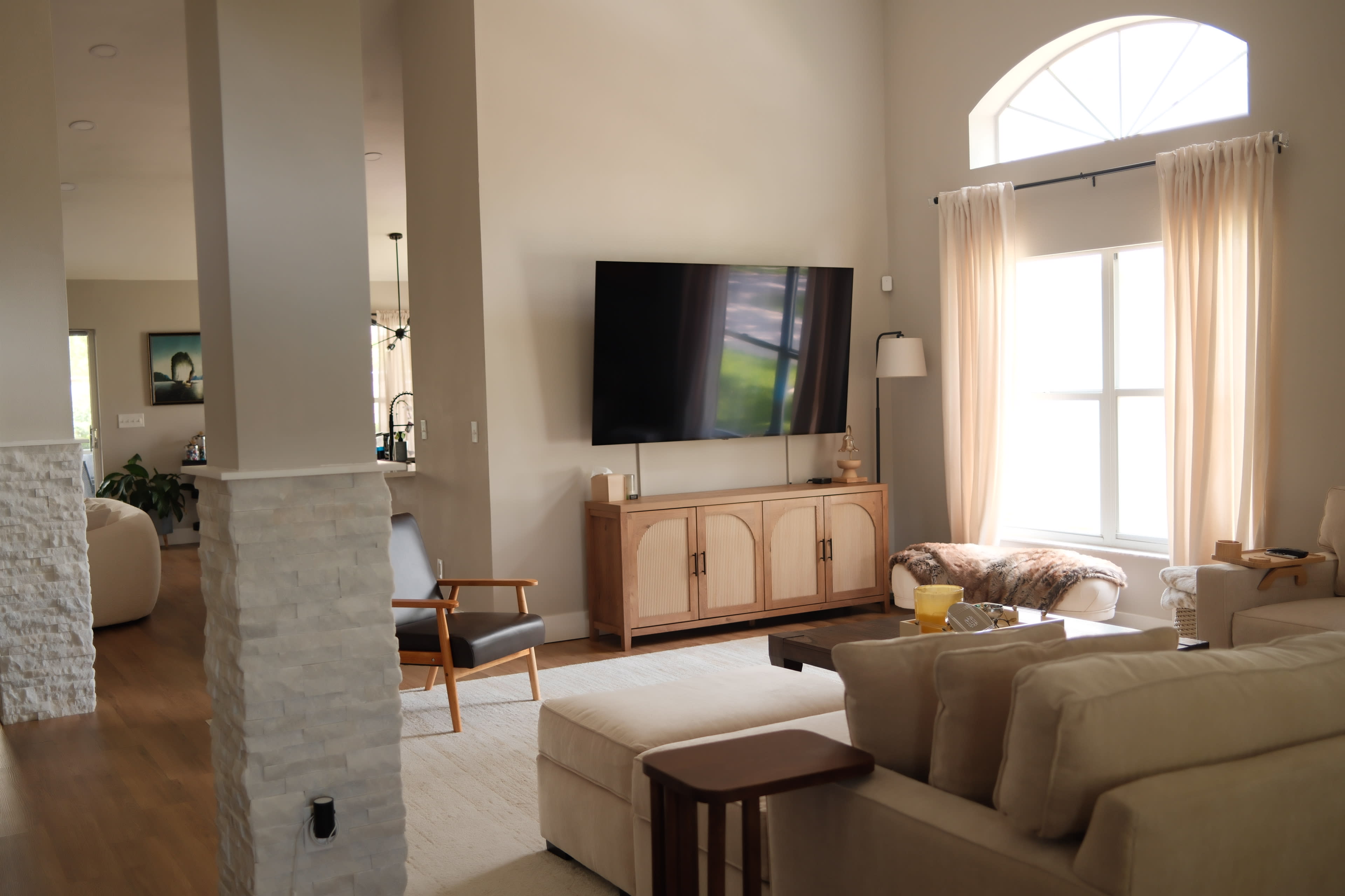 A modern living room features a large wall-mounted television, a light-colored sectional sofa, and a wooden cabinet, with natural light streaming through a bright window.