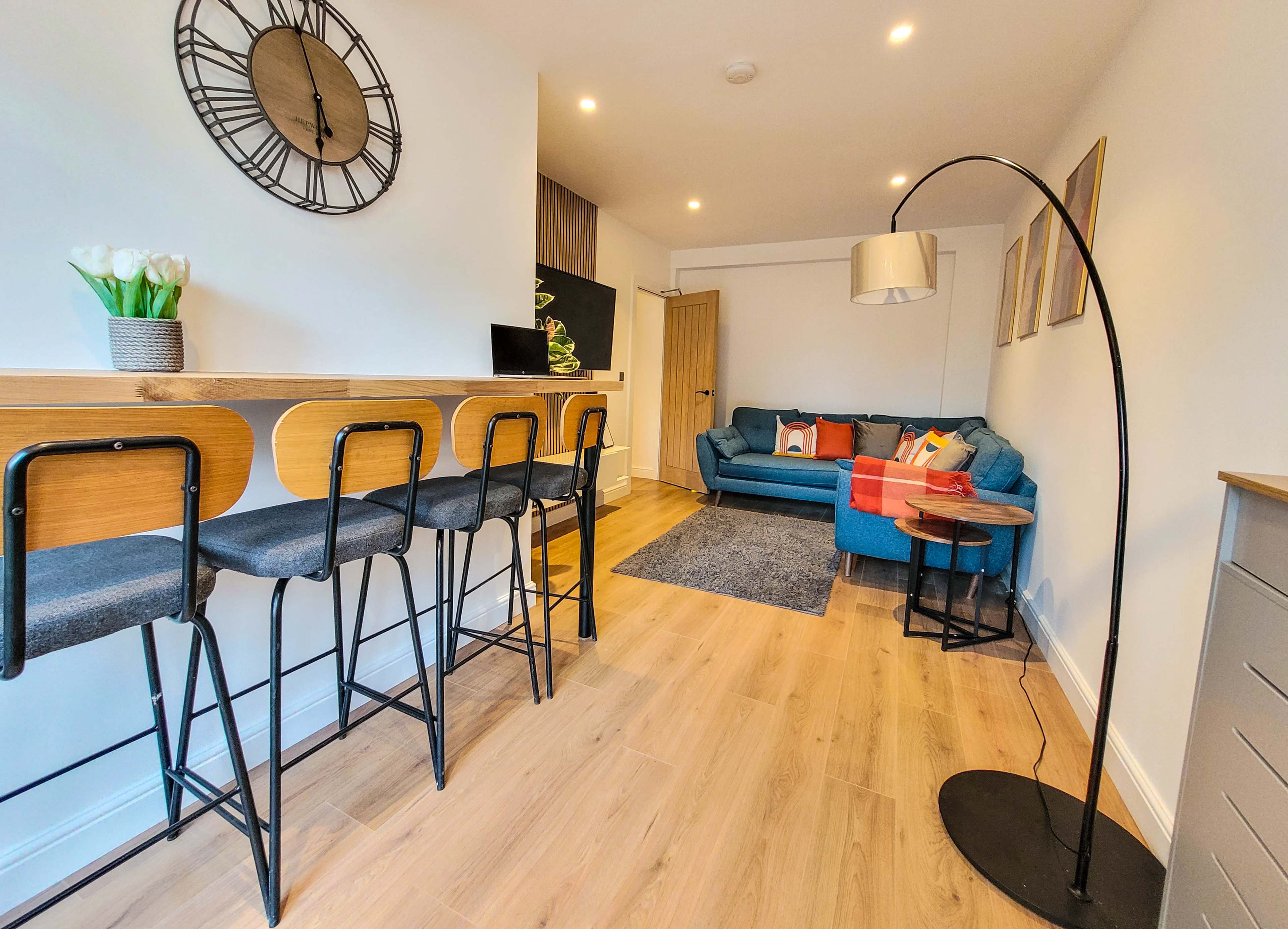 A modern open-plan living area featuring a seating arrangement with a blue couch, a small dining bar with stools, and a floor lamp.