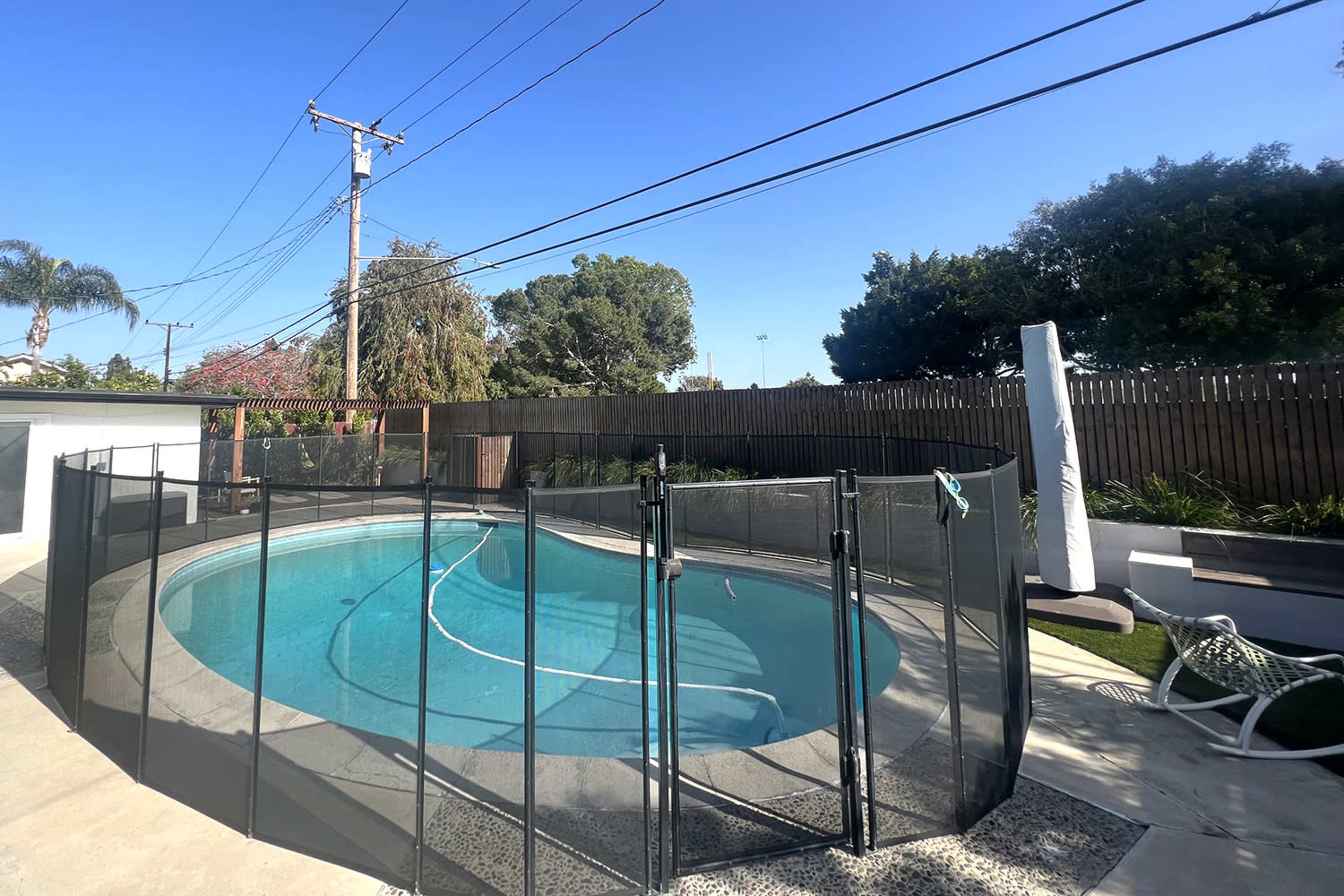 A swimming pool surrounded by a black safety fence, with a clear blue sky overhead and green foliage in the background.