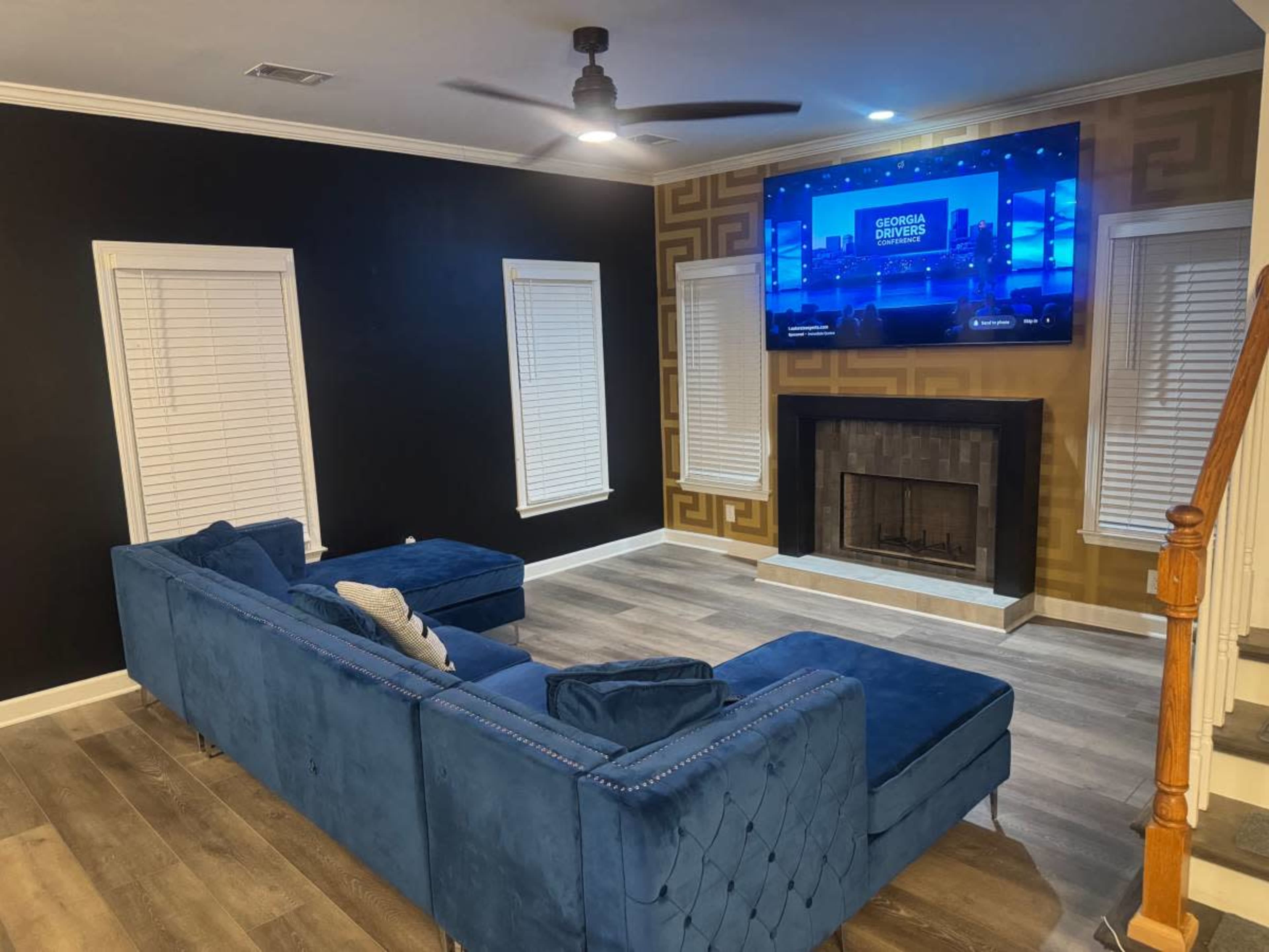The image shows a modern living room with a blue sectional sofa, a wall-mounted television displaying a program, and a fireplace beneath the TV, with additional wooden stairs visible on the right side.