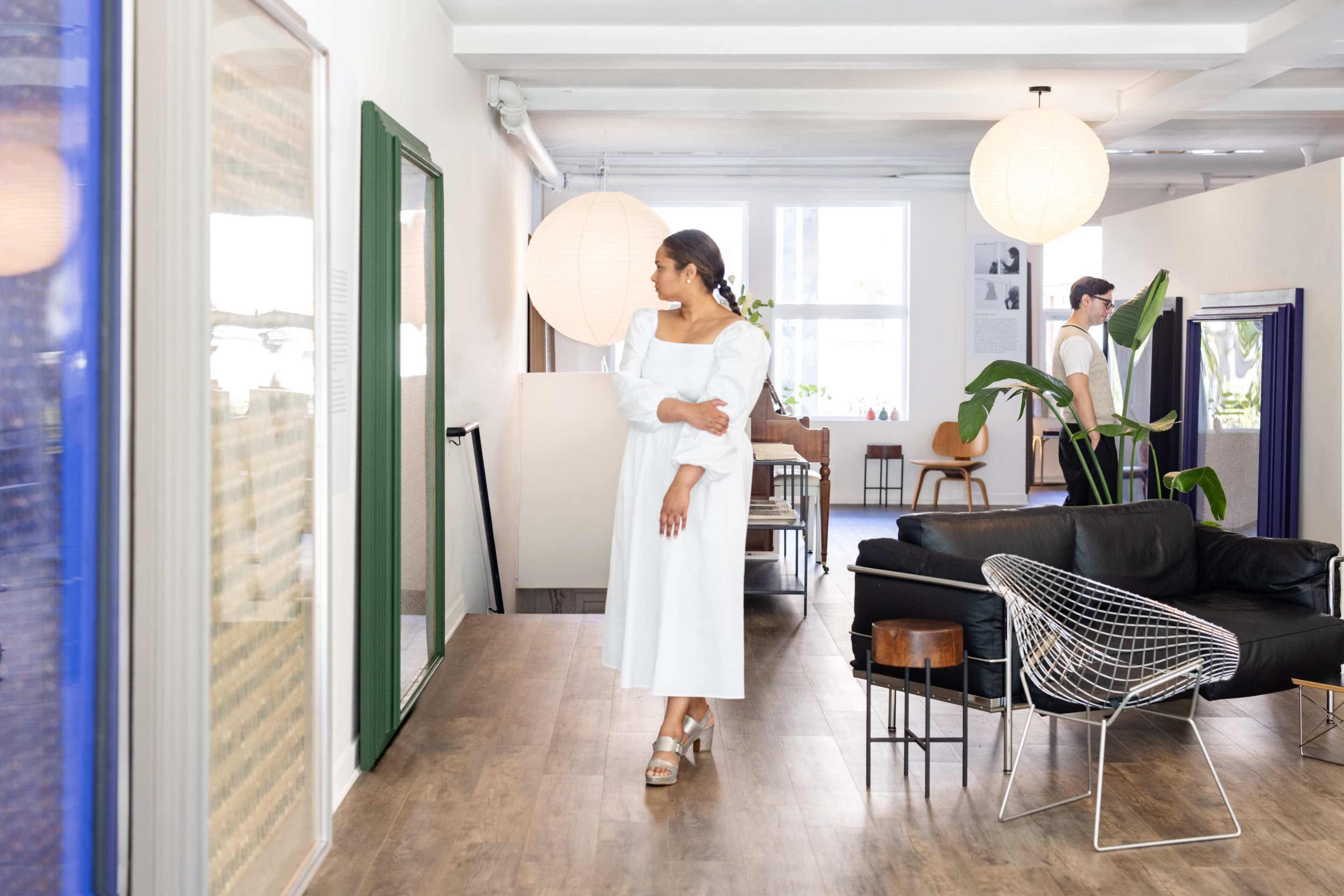 A woman in a white dress stands in a modern, airy interior space with wooden flooring and stylish furniture, while a man is visible in the background.