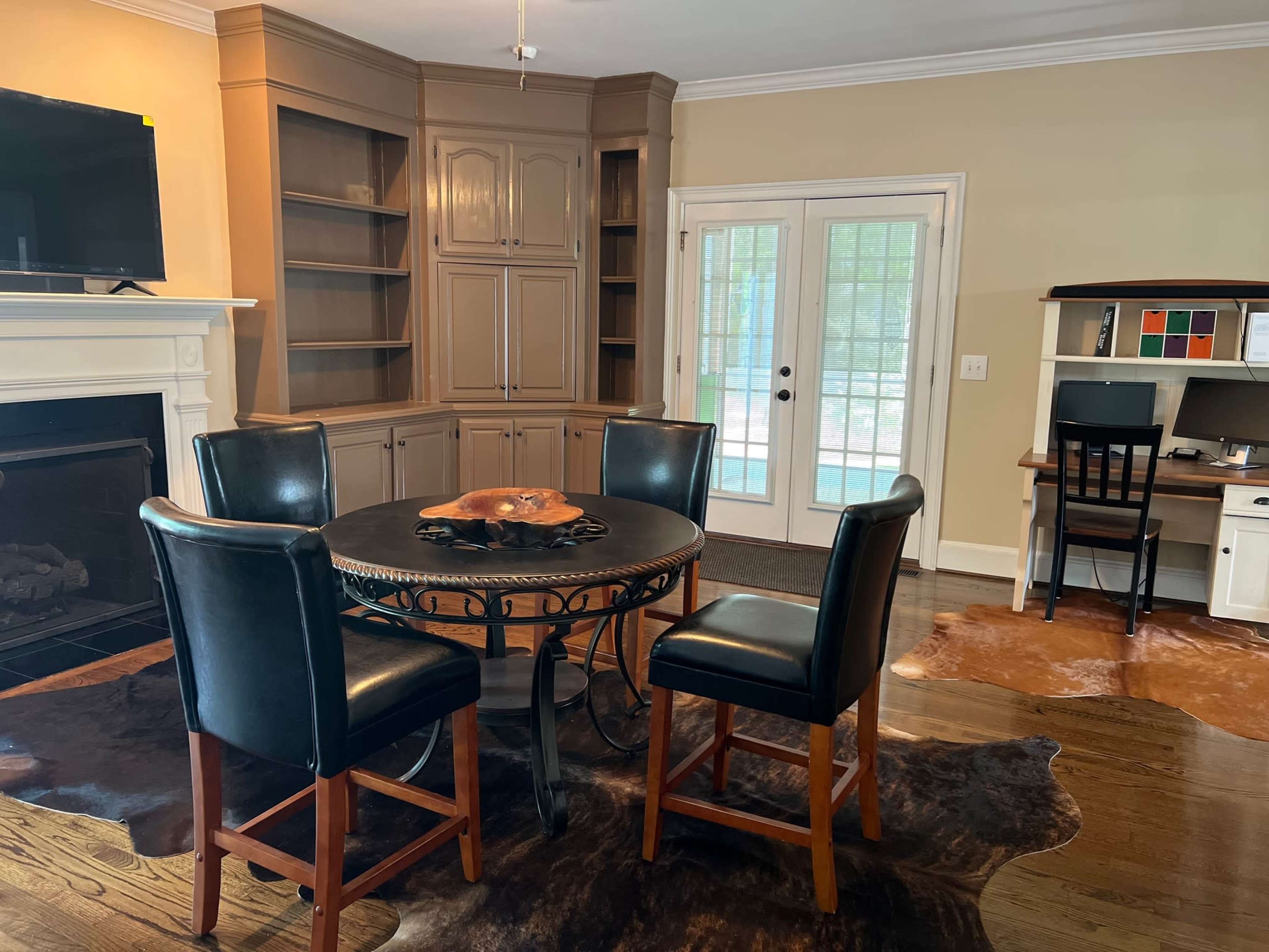 A round table with four black chairs is positioned in a room with a fireplace and built-in cabinets, along with a desk in the corner near a set of French doors.