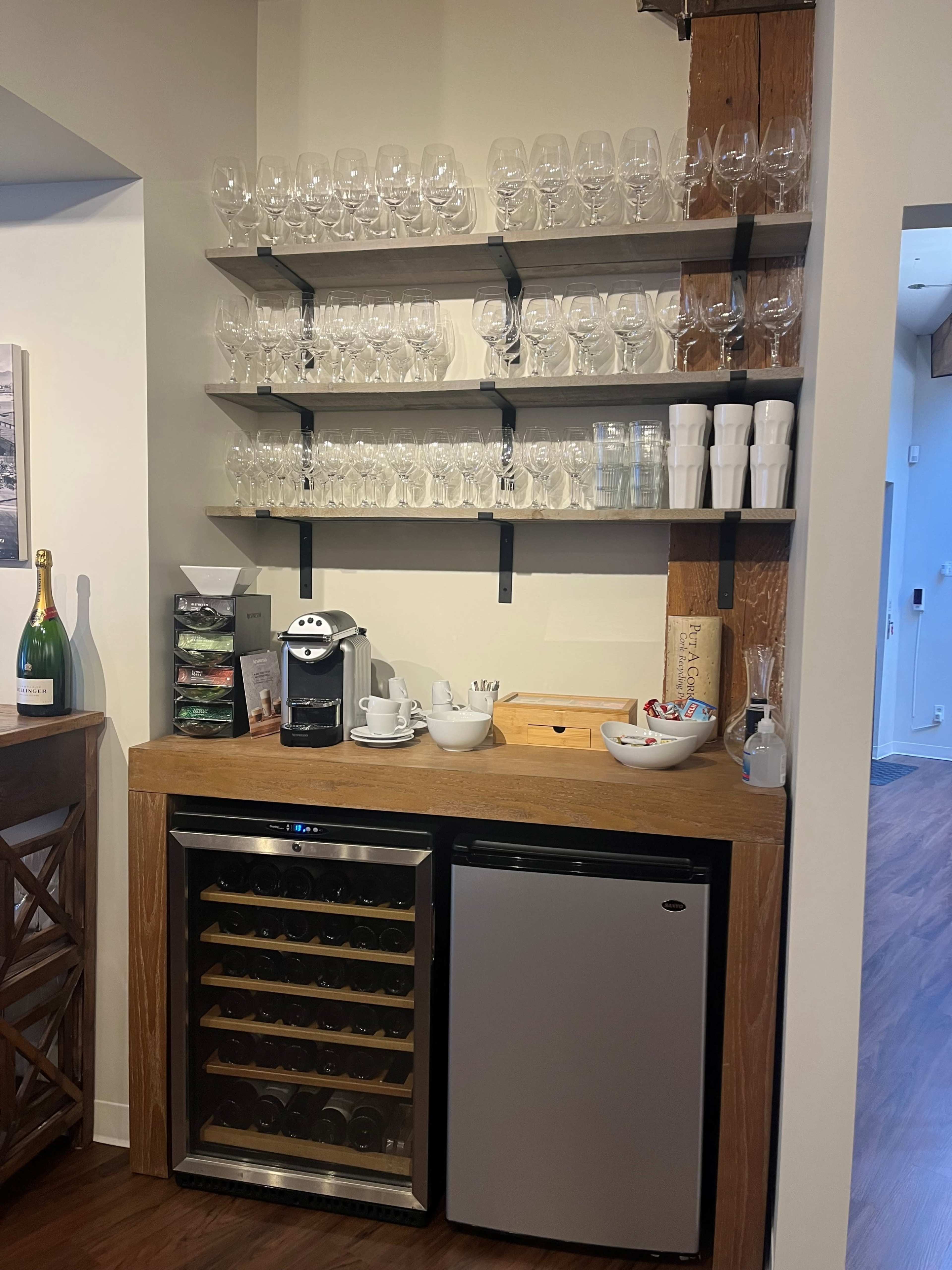 A wooden bar area with stacked glassware on shelves above a countertop featuring a coffee machine, dishware, and a small wine fridge.