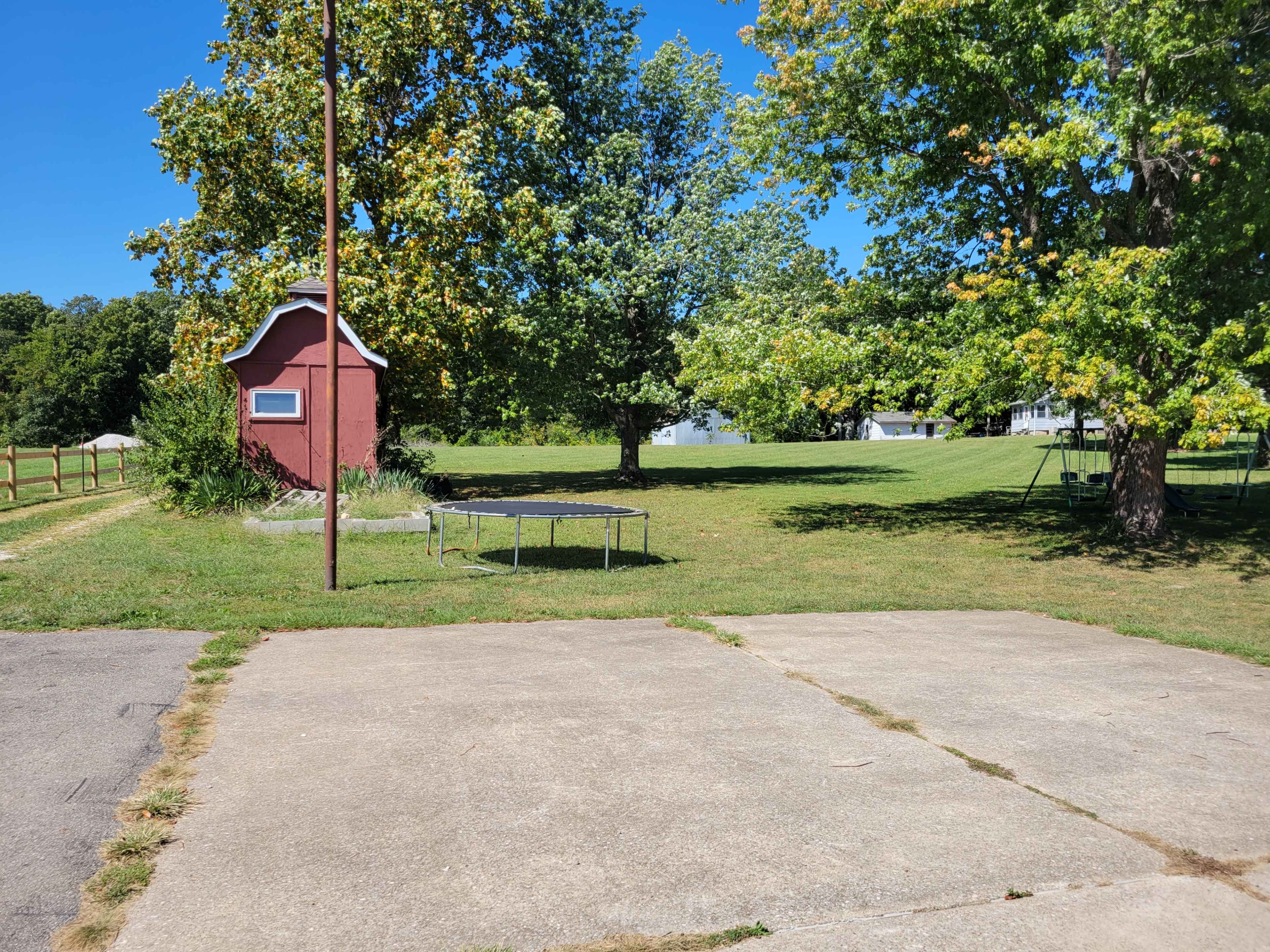 The image shows a grassy yard with a red shed, a trampoline, and a large tree, set against a clear blue sky.