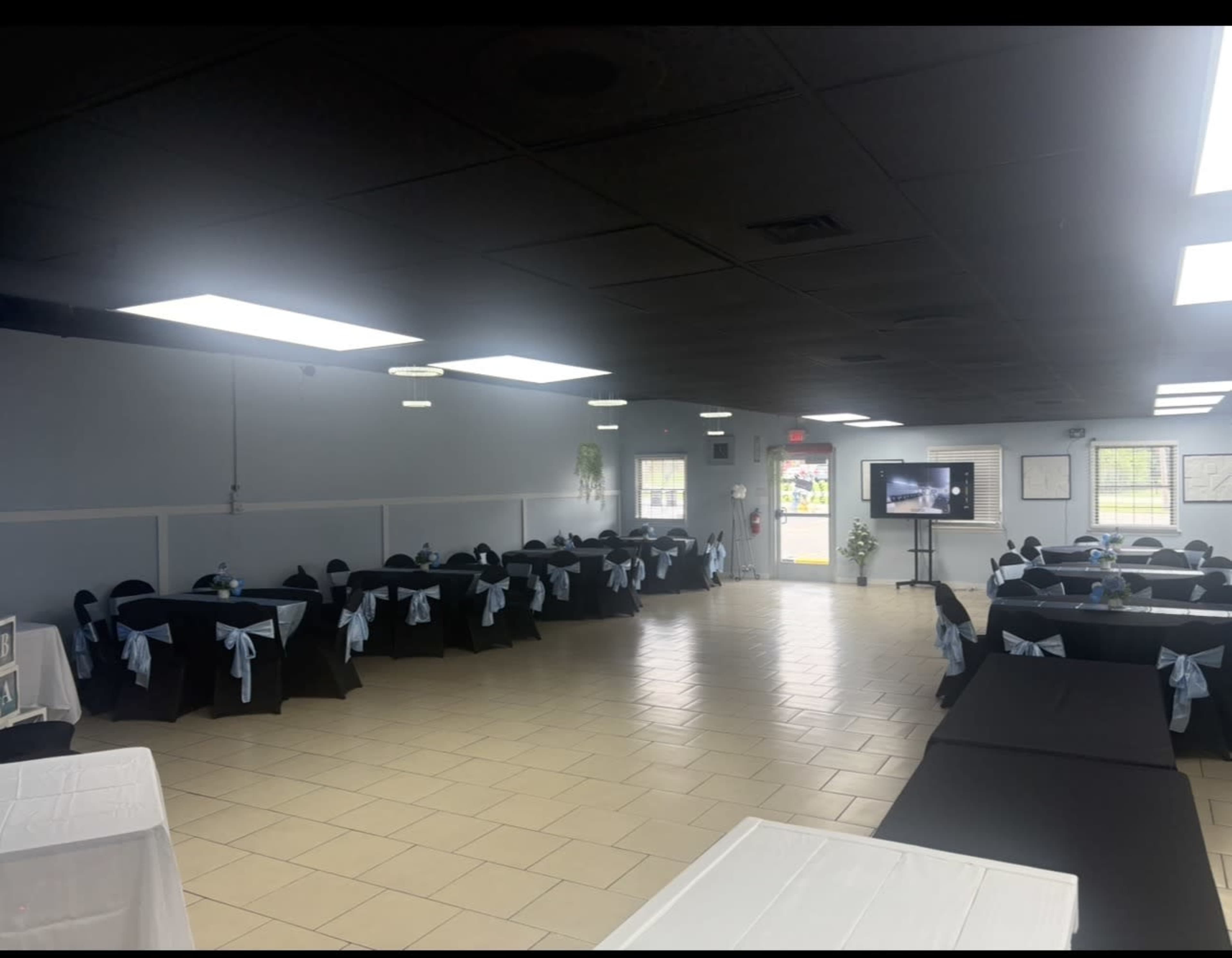 The image shows a spacious event room with several tables covered in black cloth and adorned with light blue sashes, along with a wall-mounted screen displaying a presentation in the background.