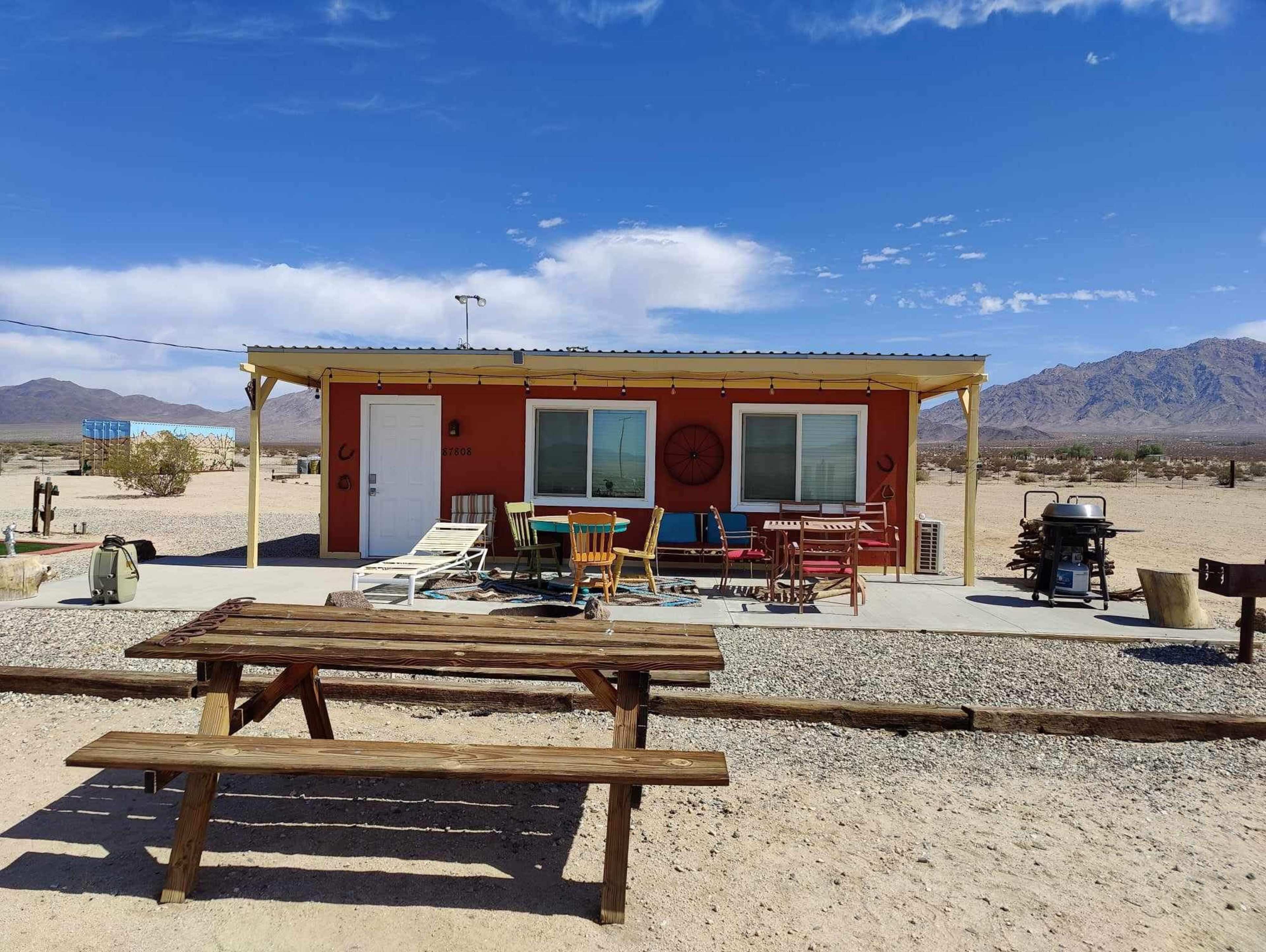 A colorful cabin with a front porch and outdoor seating is situated in a desert landscape with mountains in the background.