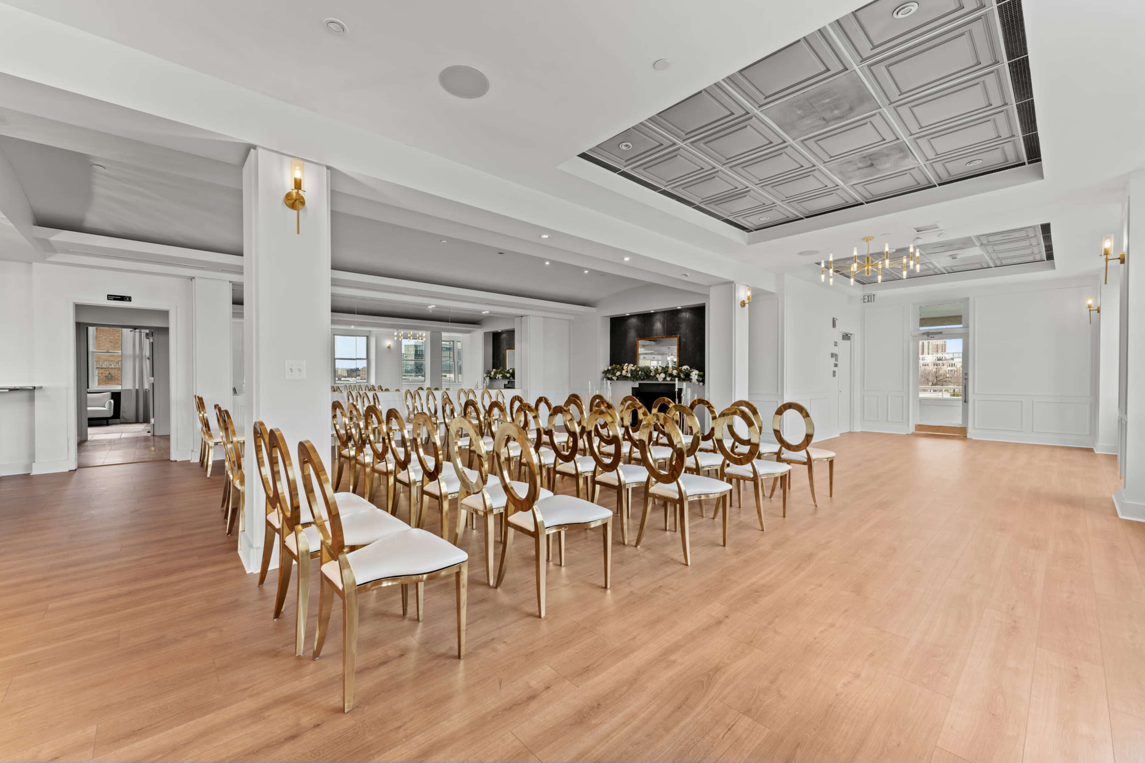 The image shows a spacious, elegantly designed room set up with rows of gold-accented chairs facing a central area, featuring large windows and a decorative ceiling.