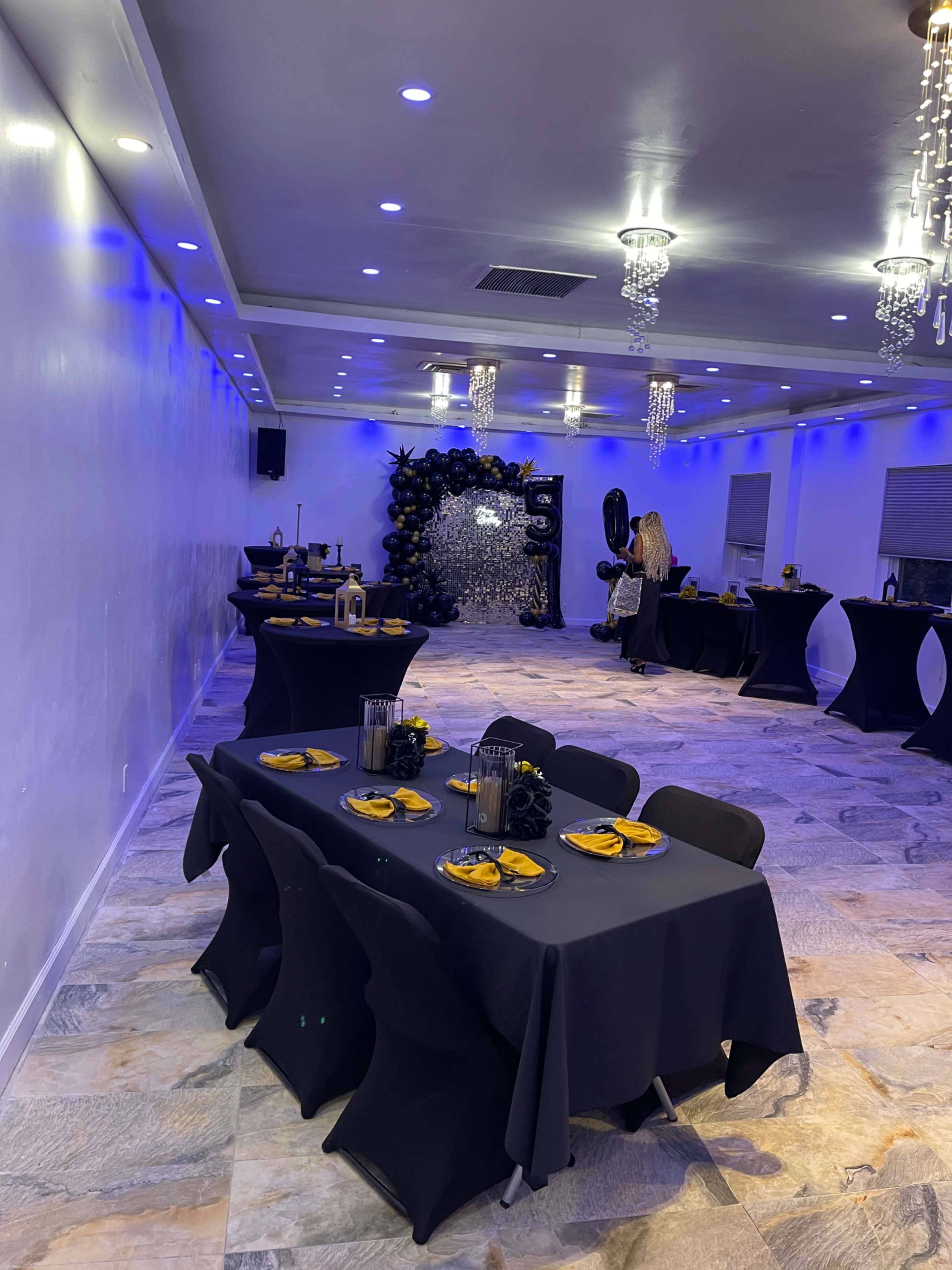 The image shows a decorated event space with black tables set for dining, blue lighting, and a backdrop featuring balloons and decorations.