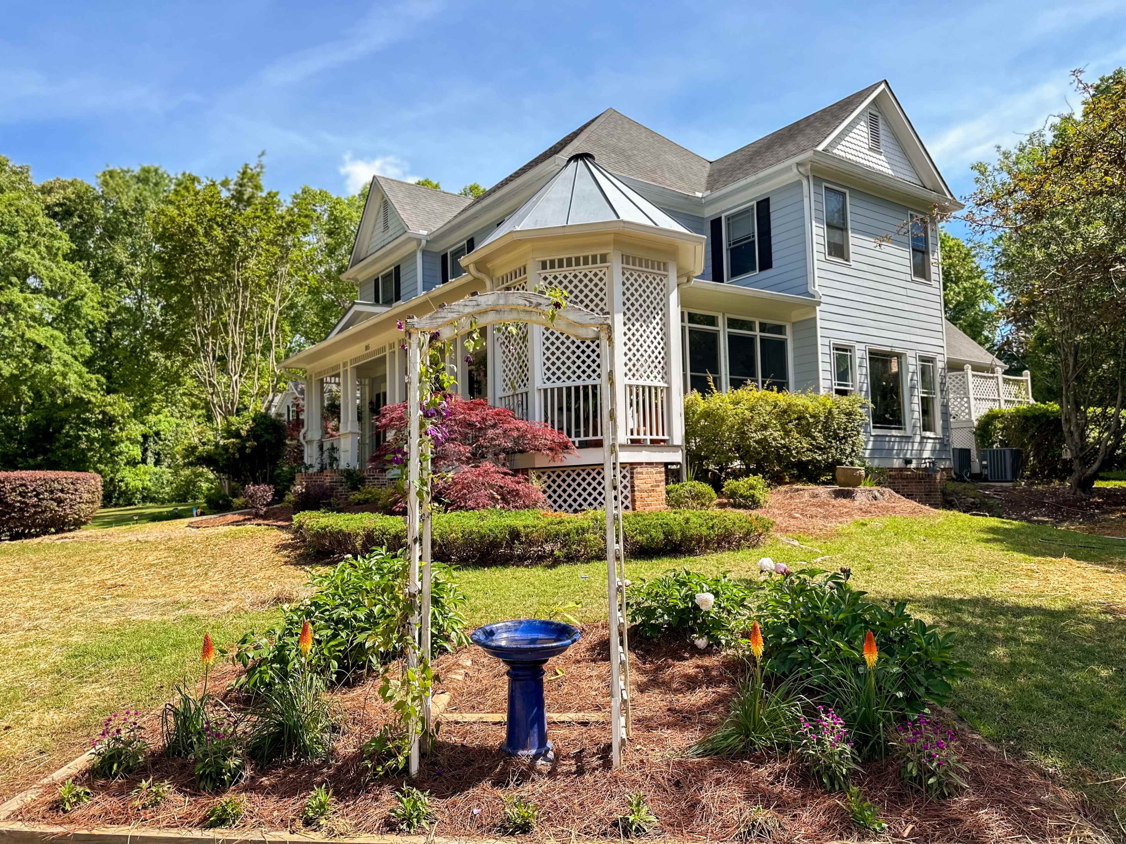 A two-story blue house with a front porch and garden features a white trellis and a birdbath surrounded by flowers.