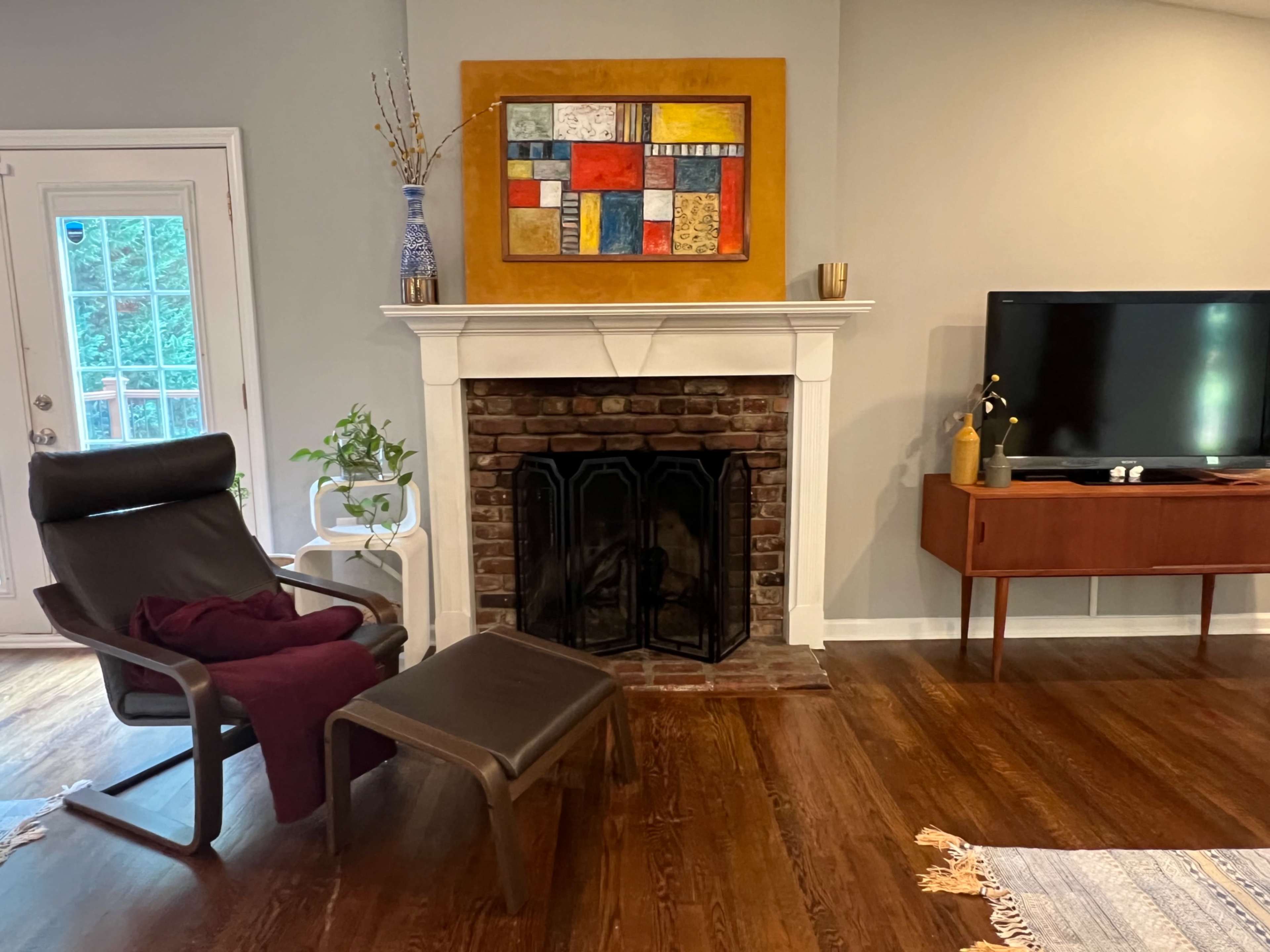 The image shows a living room featuring a brick fireplace, a modern armchair with a blanket, a television on a wooden stand, and a colorful abstract painting above the fireplace.