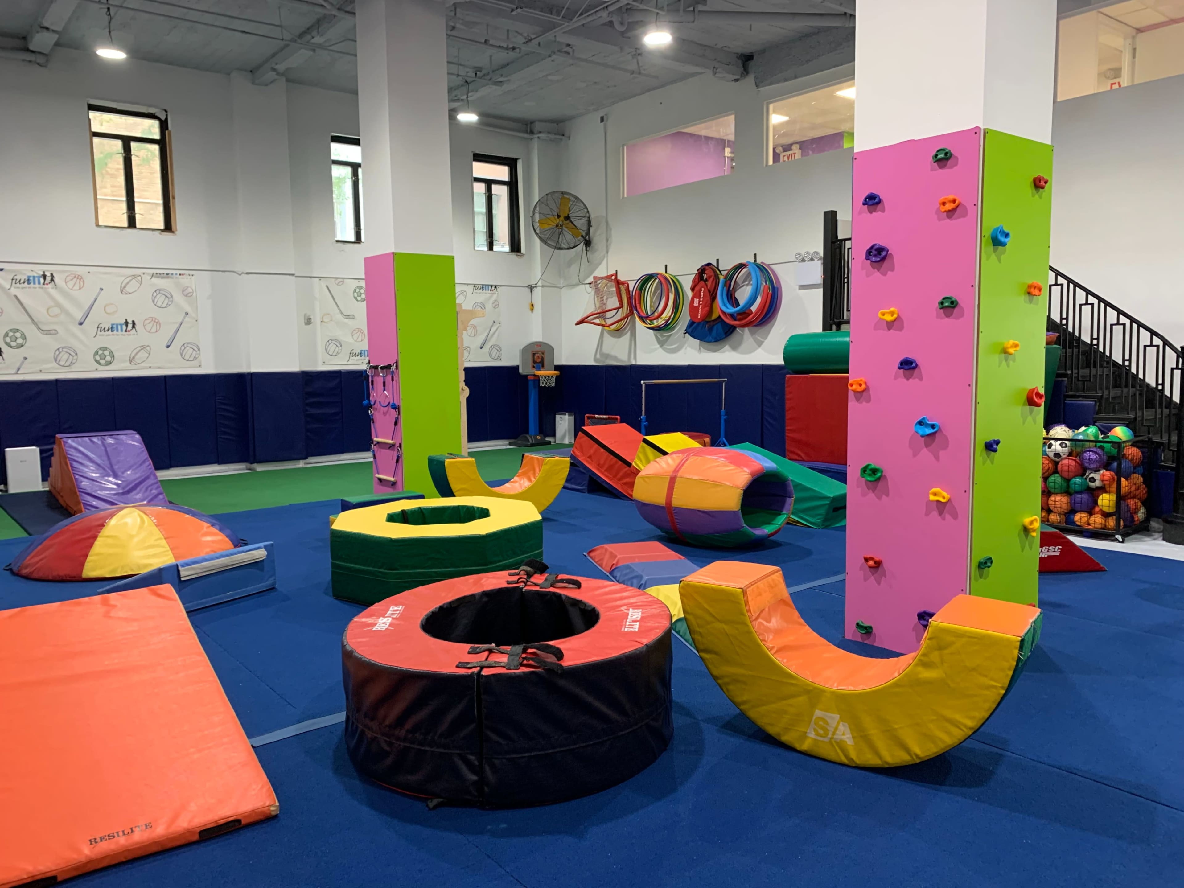 The image shows an indoor play area filled with colorful gymnastic equipment, including foam shapes, climbing walls, and mats on a blue floor.