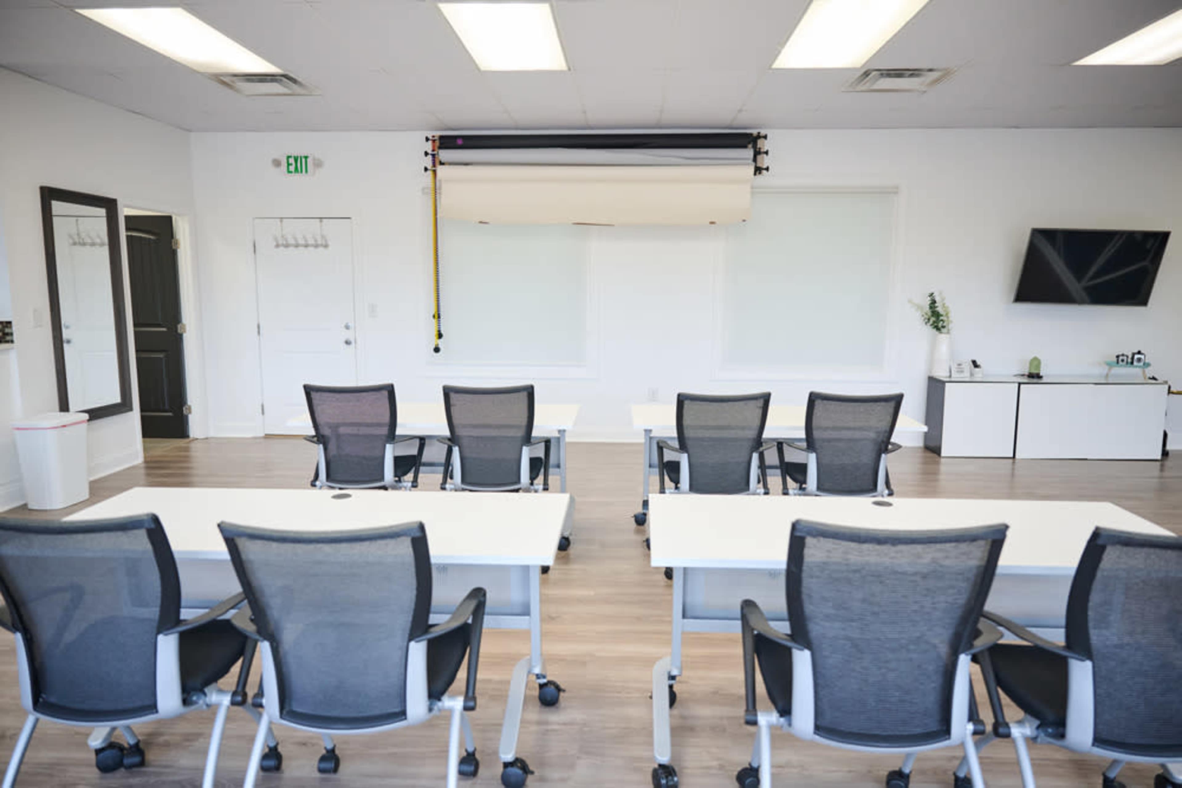 A modern conference room features several white meeting tables paired with mesh office chairs, all arranged facing a blank wall with a mounted screen and a television.